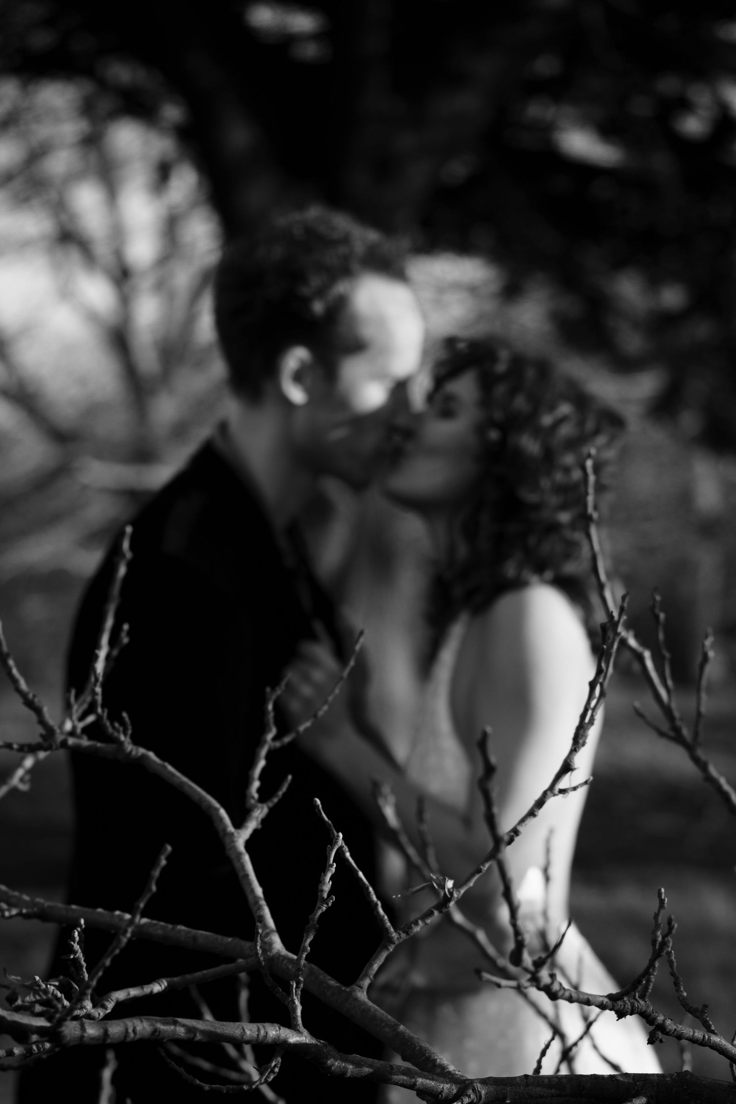 A black and white artistic photo of a couple embracing outdoors with dramatic shadows and branches.