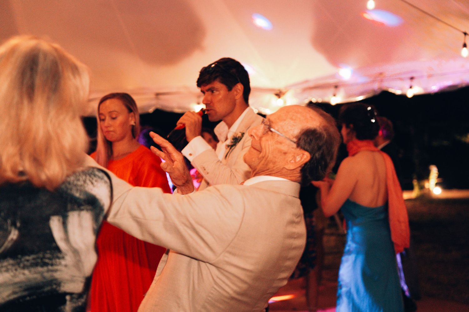 Guests dancing joyfully at an elegant outdoor wedding reception under a white tent with warm lighting.