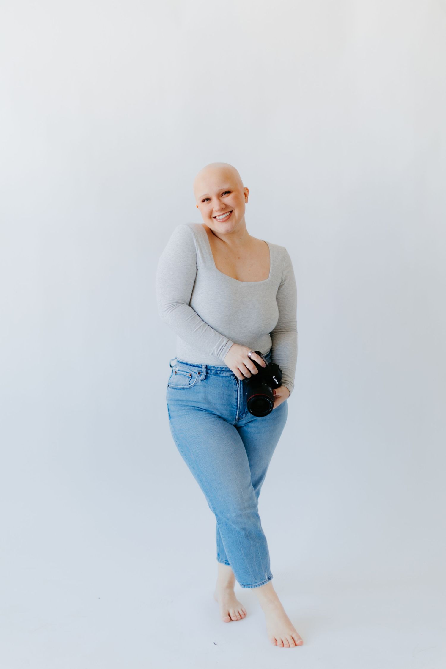 Smiling bald woman in gray top and jeans holding a camera, standing barefoot against a white background.