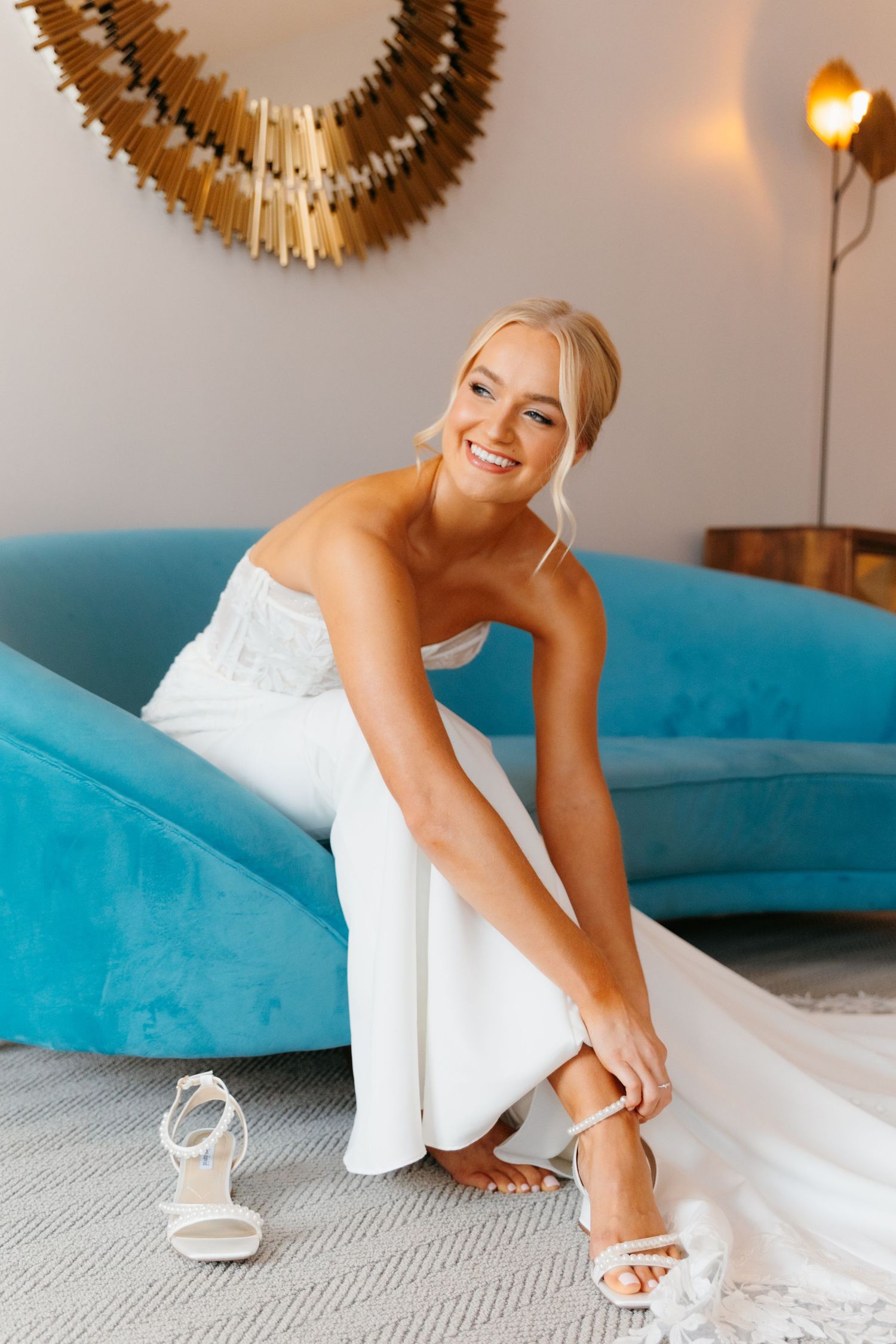 White bridal shoes being put on while sitting on a bright blue velvet couch.
