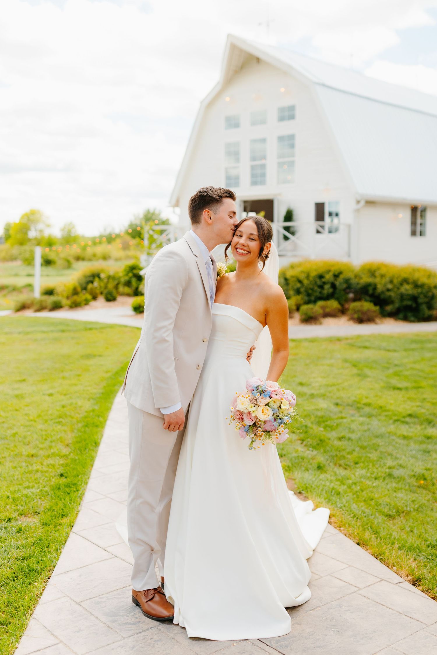 A couple in light-colored wedding attire shares tender moments outside a white farmhouse.