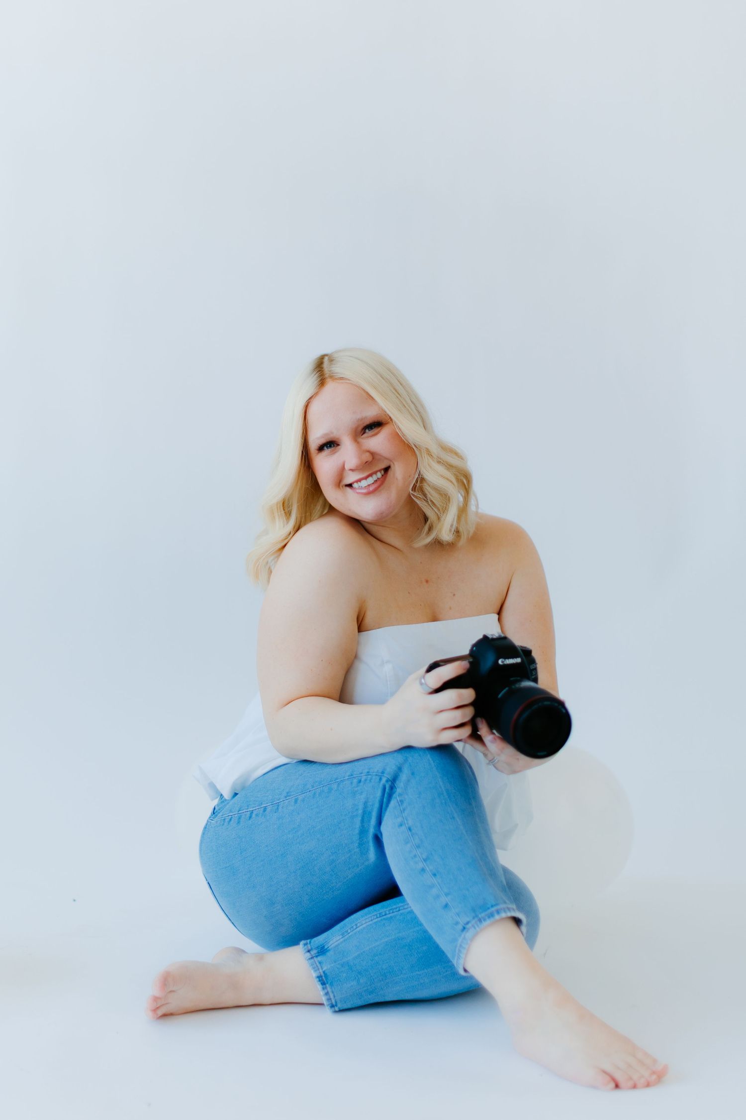 Professional photographer wearing white top and jeans holds DSLR camera while sitting on white backdrop.
