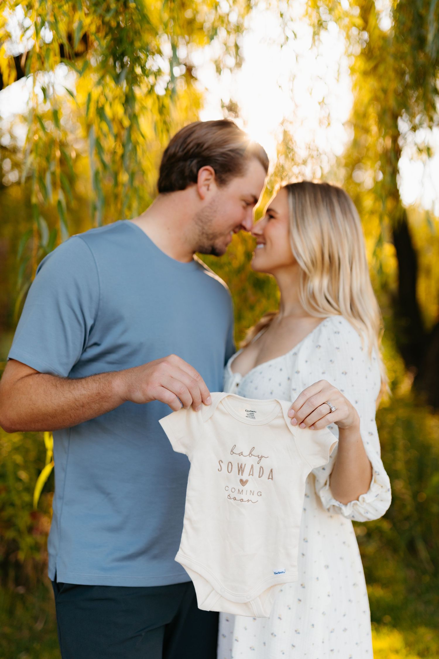 Couple holds up baby onesie together in outdoor autumn setting.