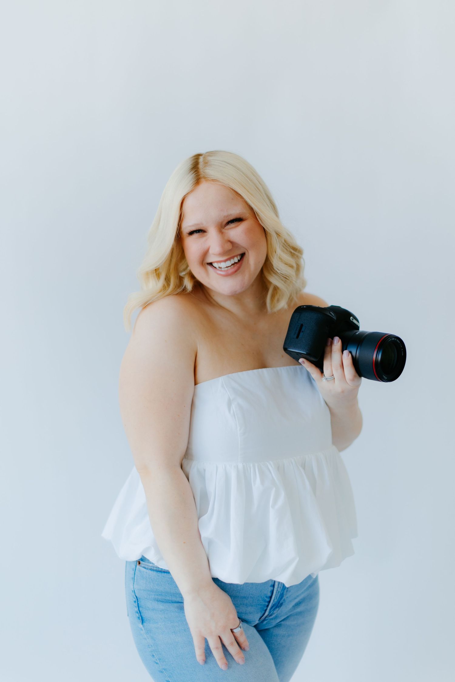 Photographer in white blouse and jeans poses with professional camera against white background.