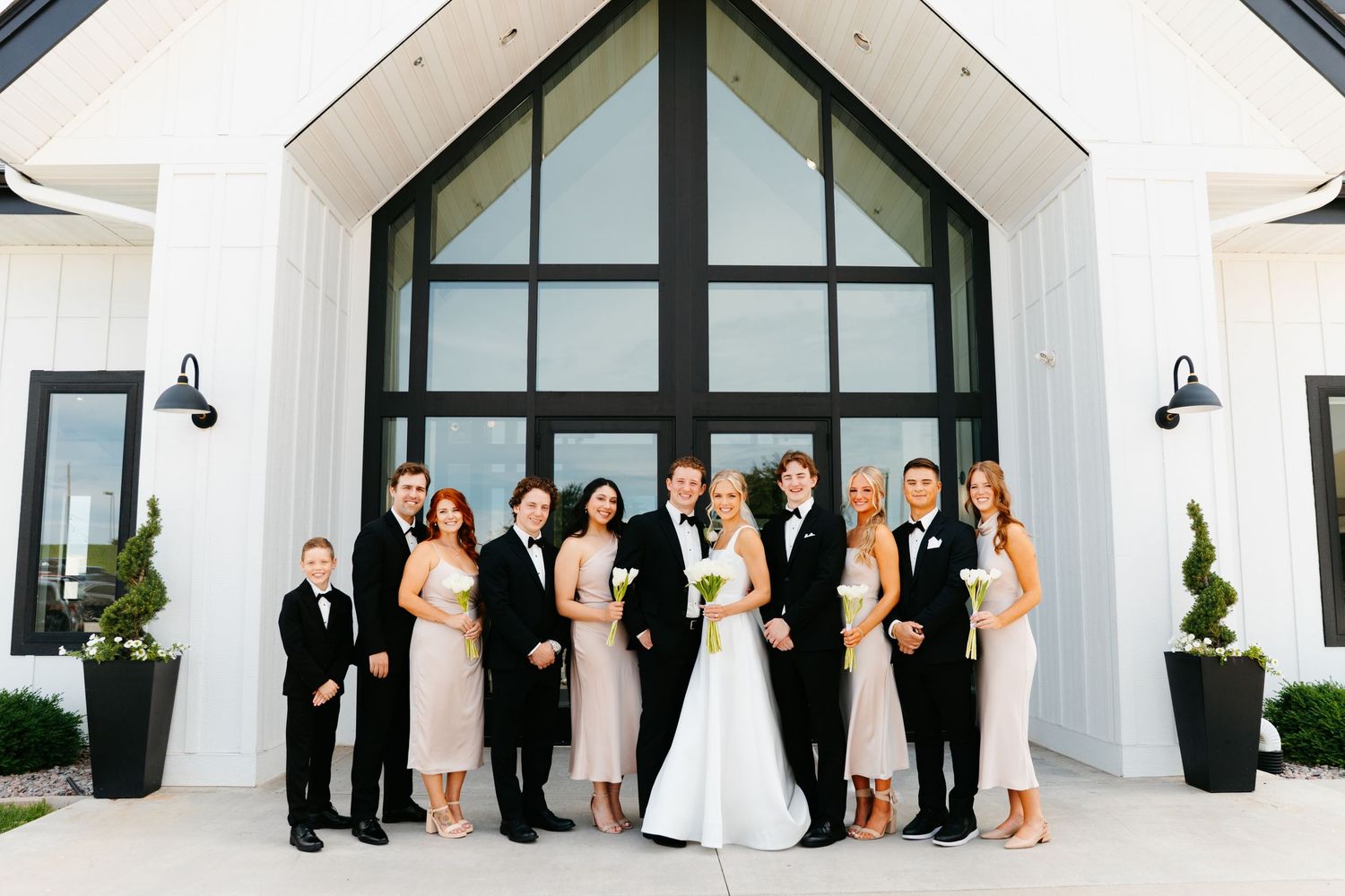 Wedding party poses in front of a modern white chapel with black-framed windows.