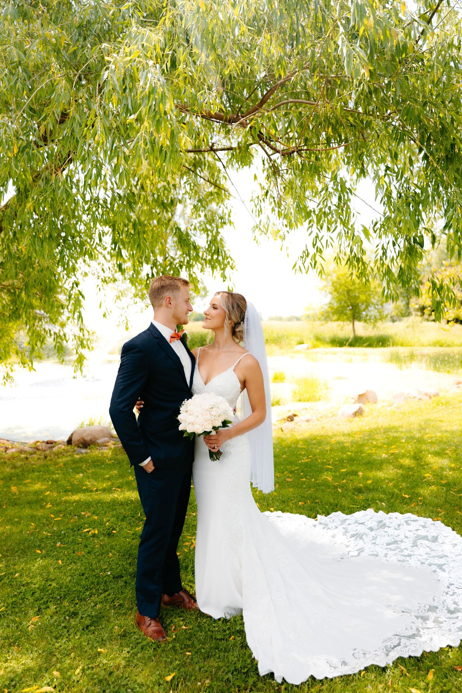Couple shares intimate moment under willow tree with flowing white wedding dress trailing on grass.