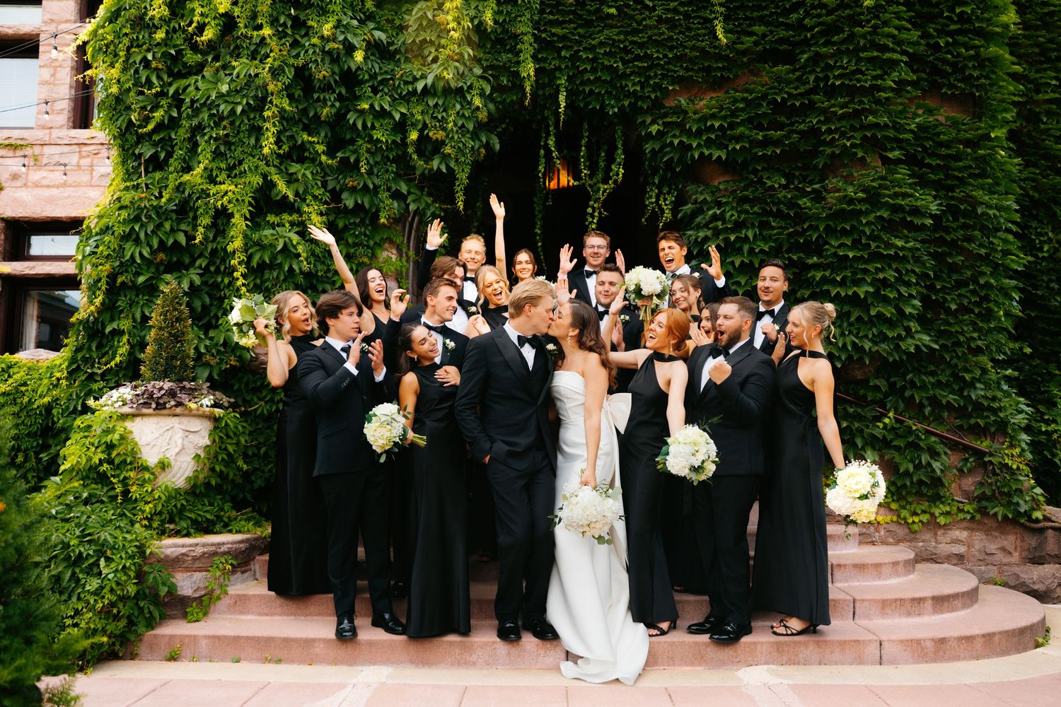 Wedding party in formal attire poses together on steps with ivy-covered walls.