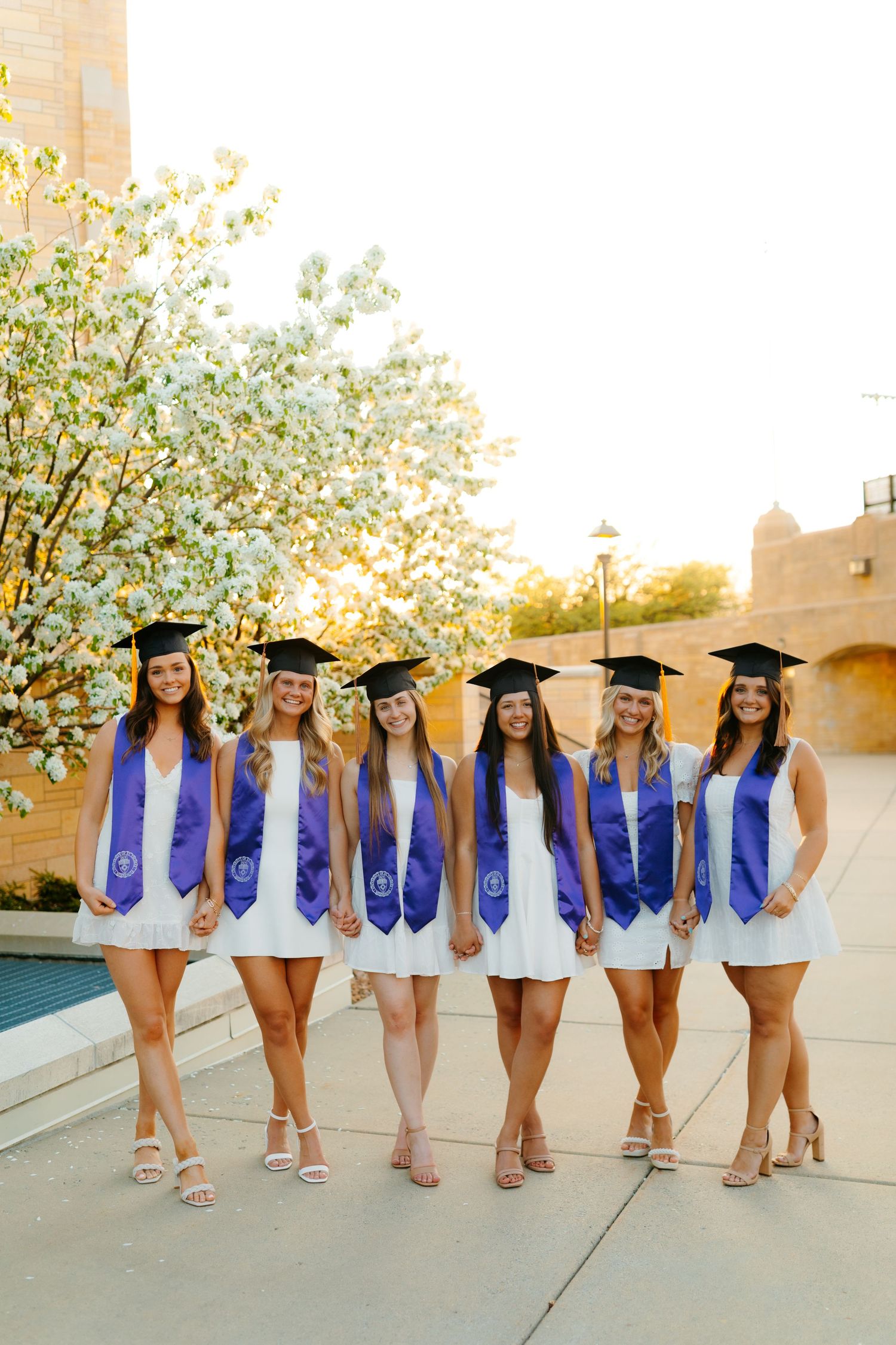 Graduates posing in white dresses and purple stoles with graduation caps.