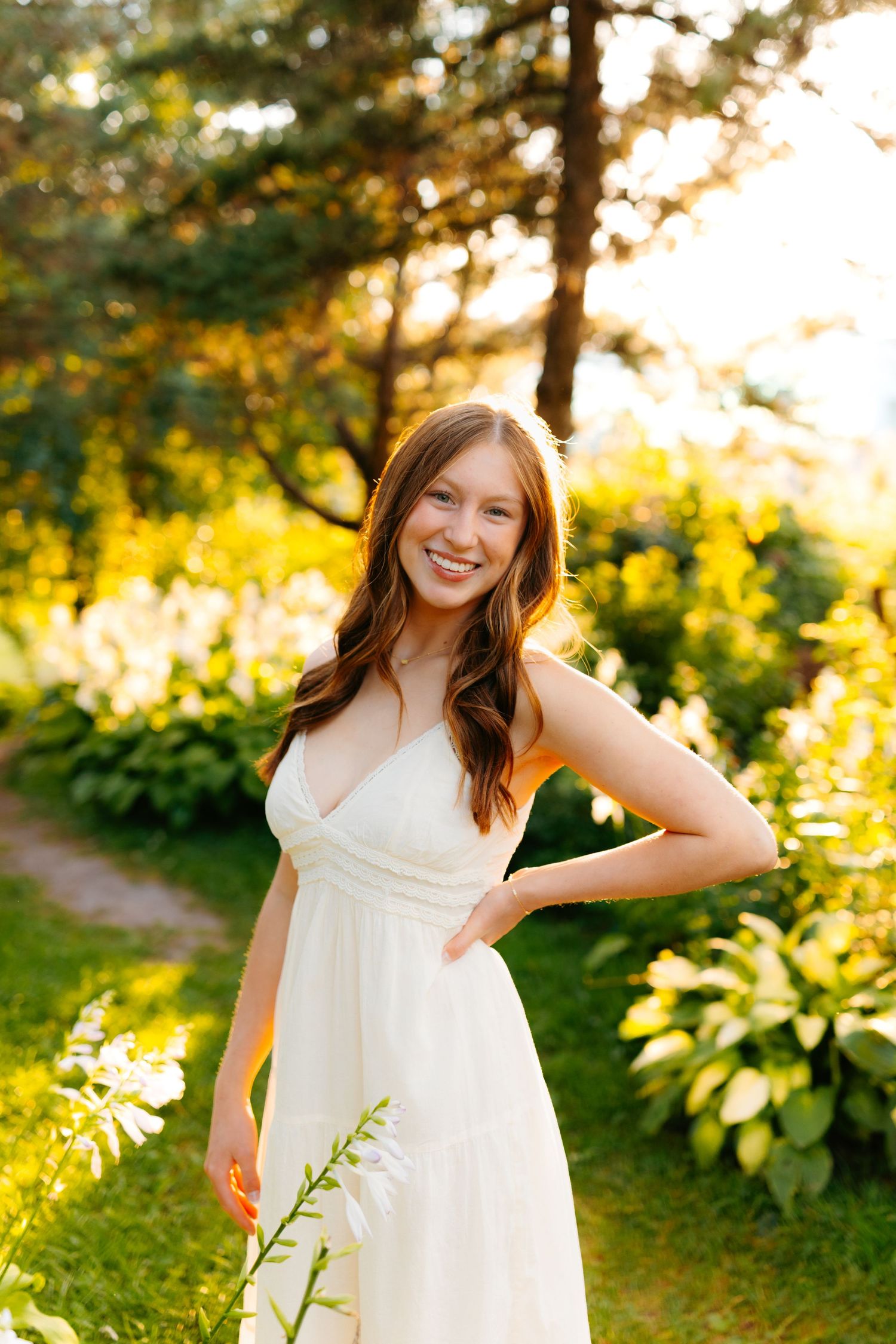 A white sundress glows in golden sunset light among lush garden foliage.
