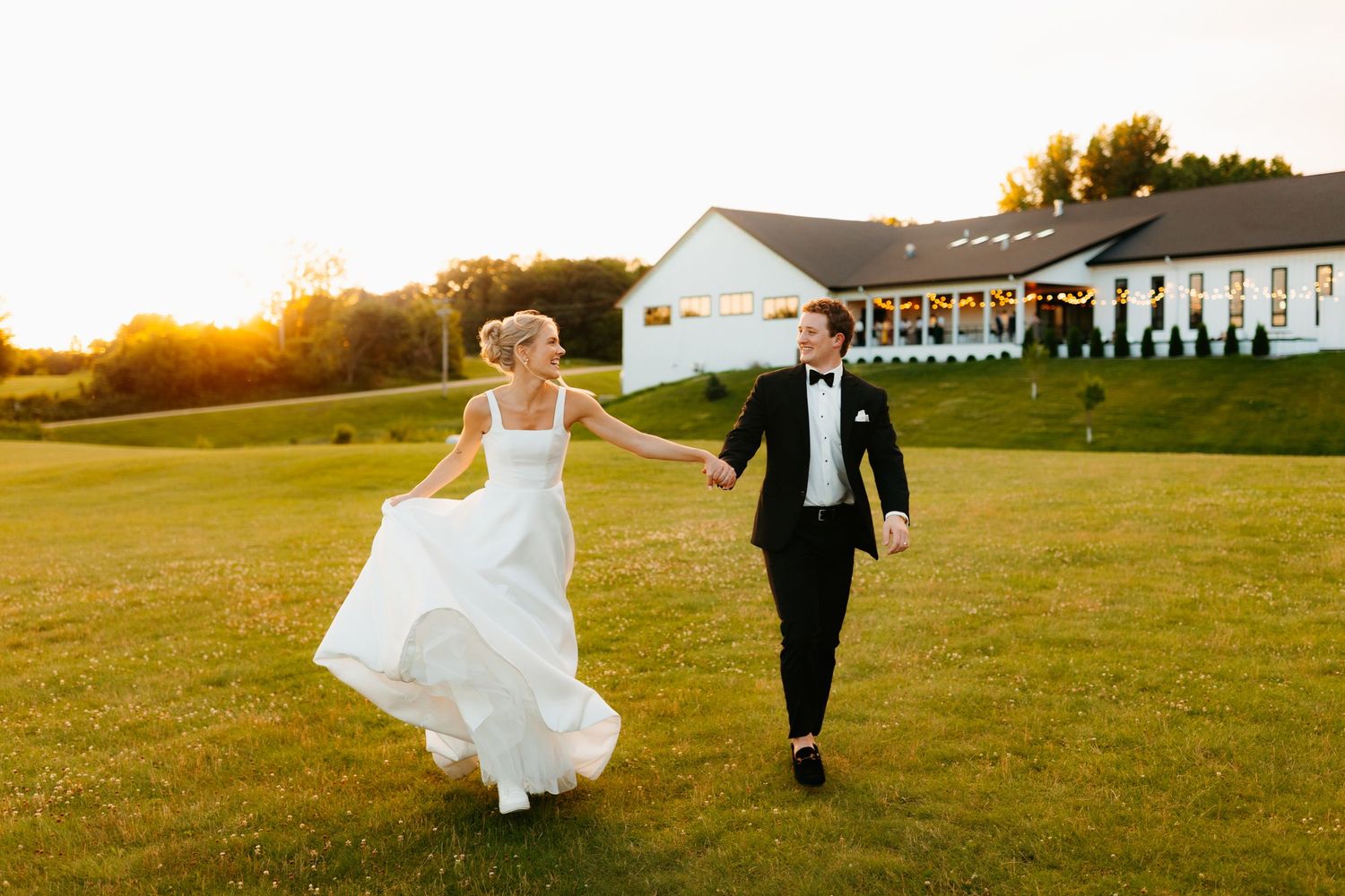 Newlyweds walk hand in hand across a grassy field at sunset with buildings in background.