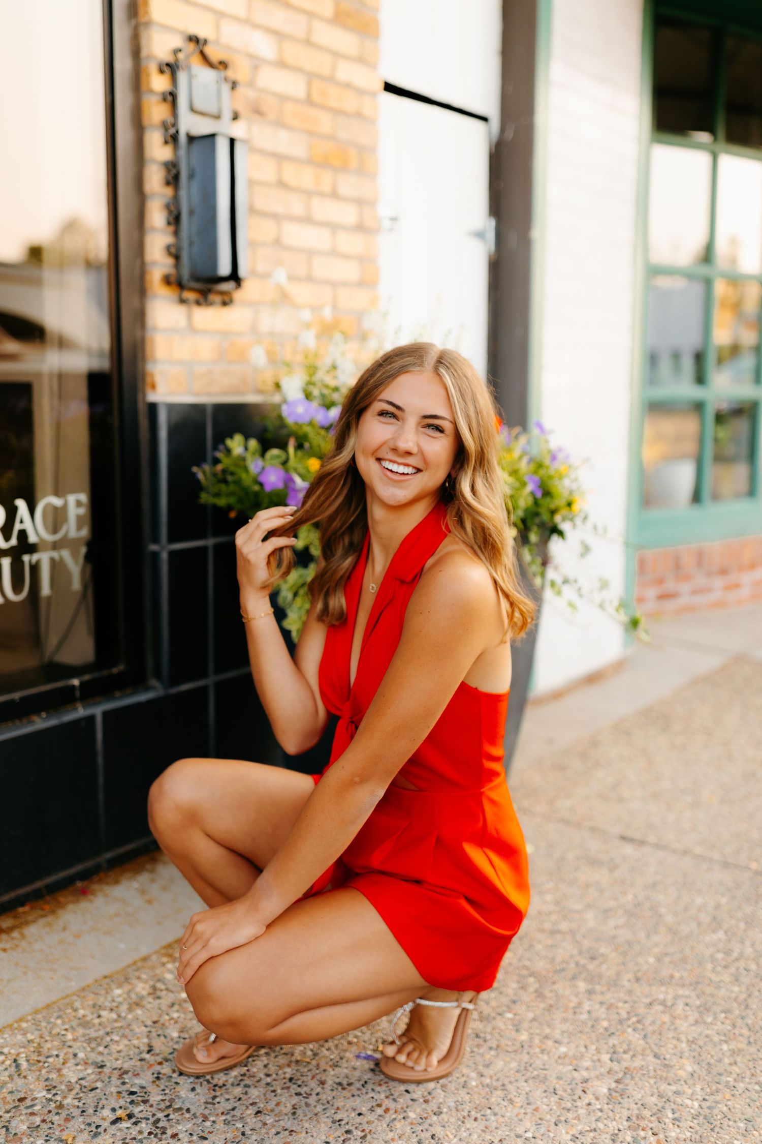 A woman in a red dress crouches and smiles outside a brick storefront.