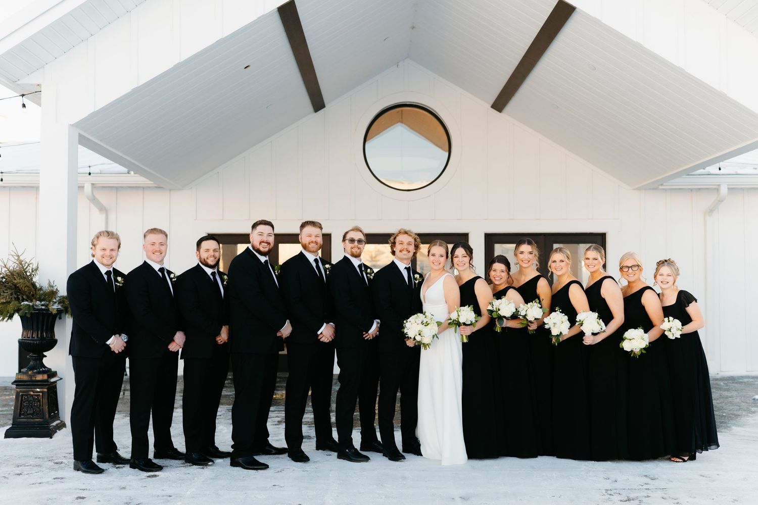 Large wedding party in black and white formal attire posed in modern white venue.
