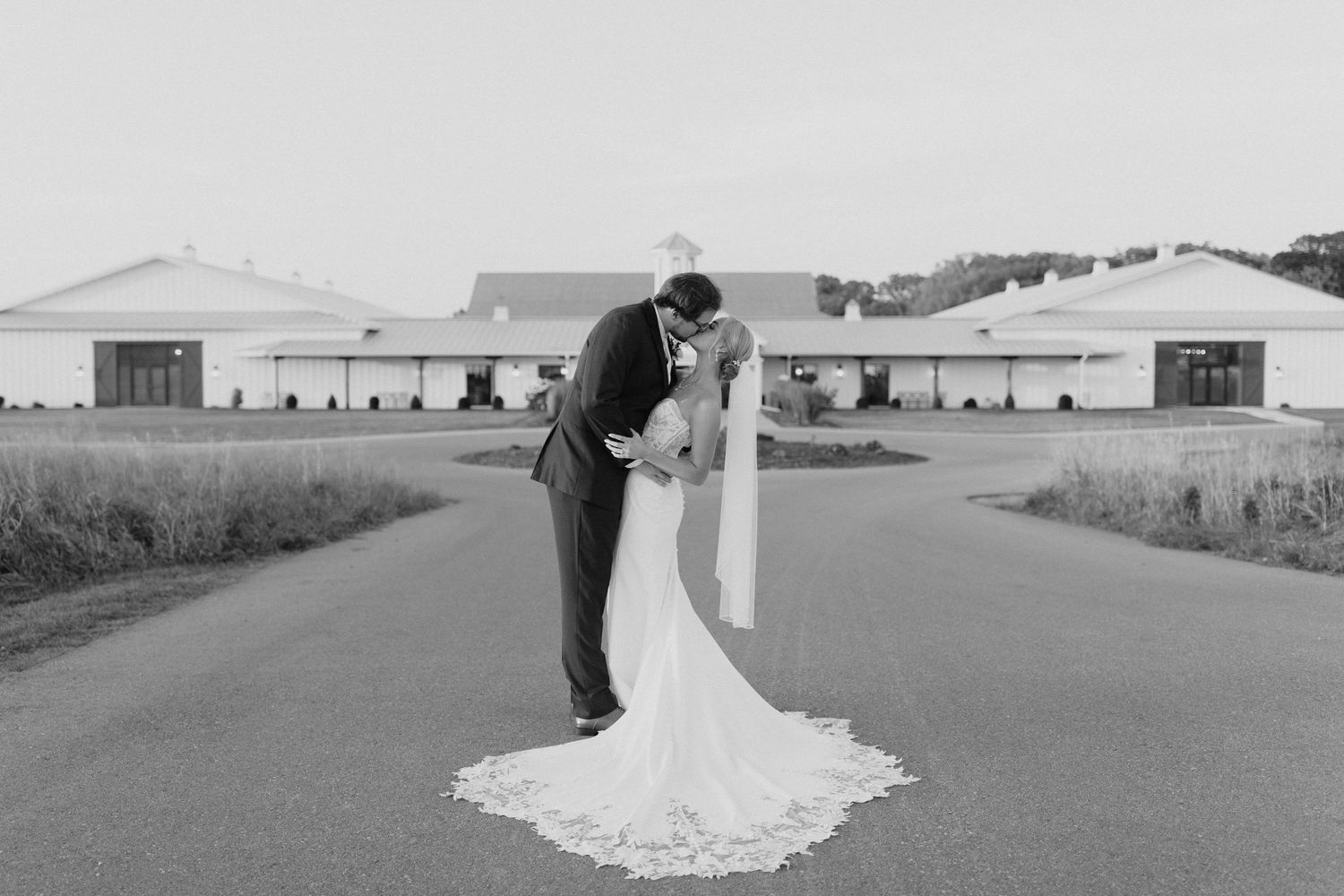 A couple in wedding attire walks together through a scenic field with mountains in the distance.