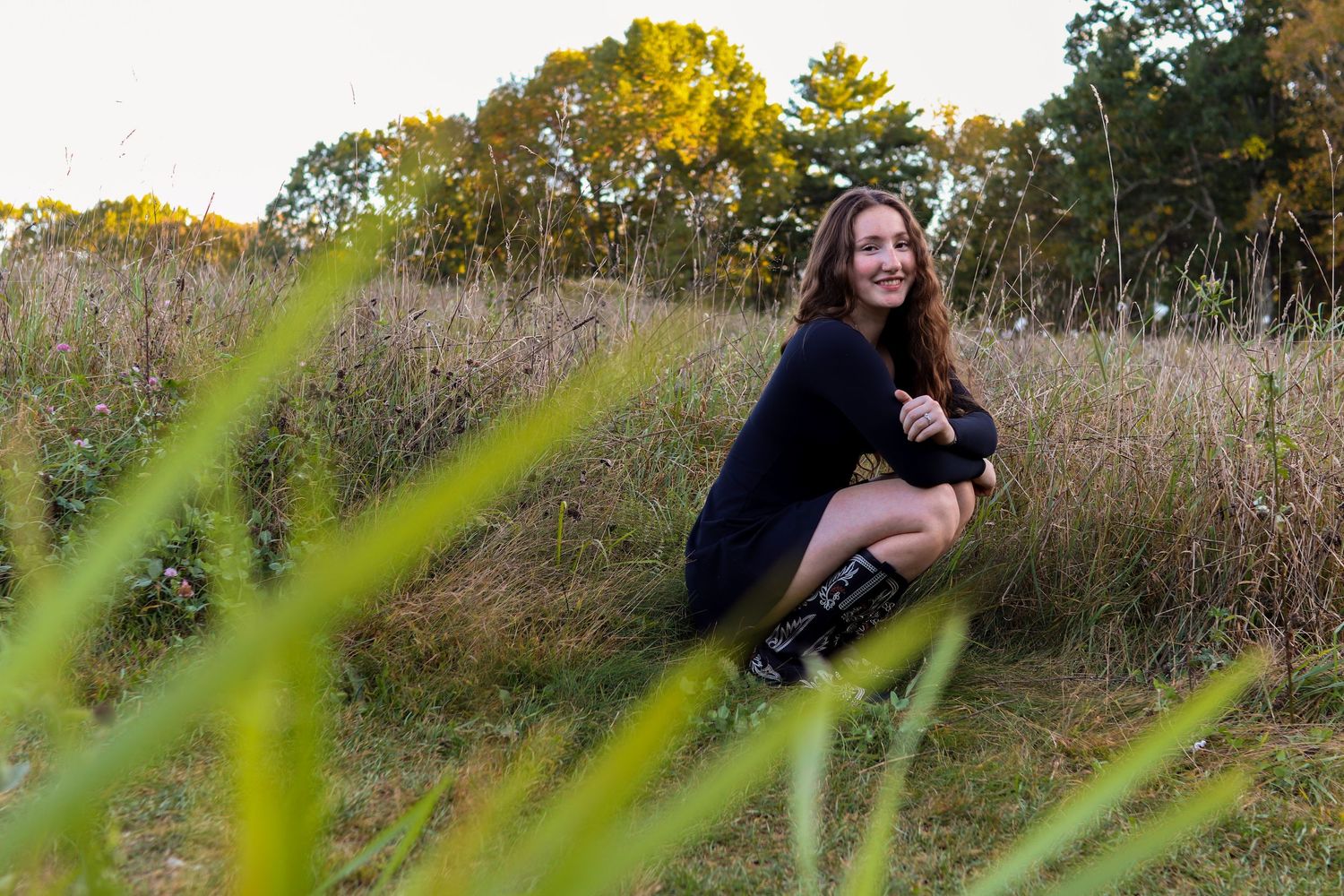 Senior photo of girl in black dress and cowboy/ western boots. In the tall grass smiling at golden hour. Central MA