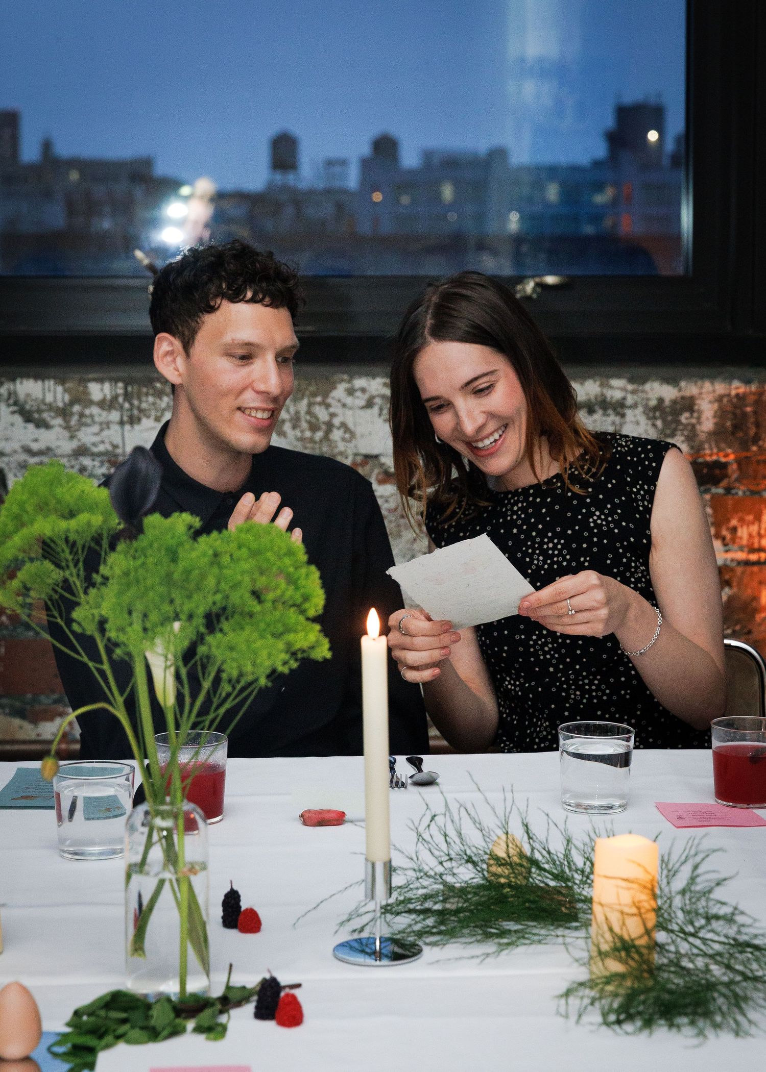A man and woman laugh together reading a card at a candlelit dinner table with floral arrangements.