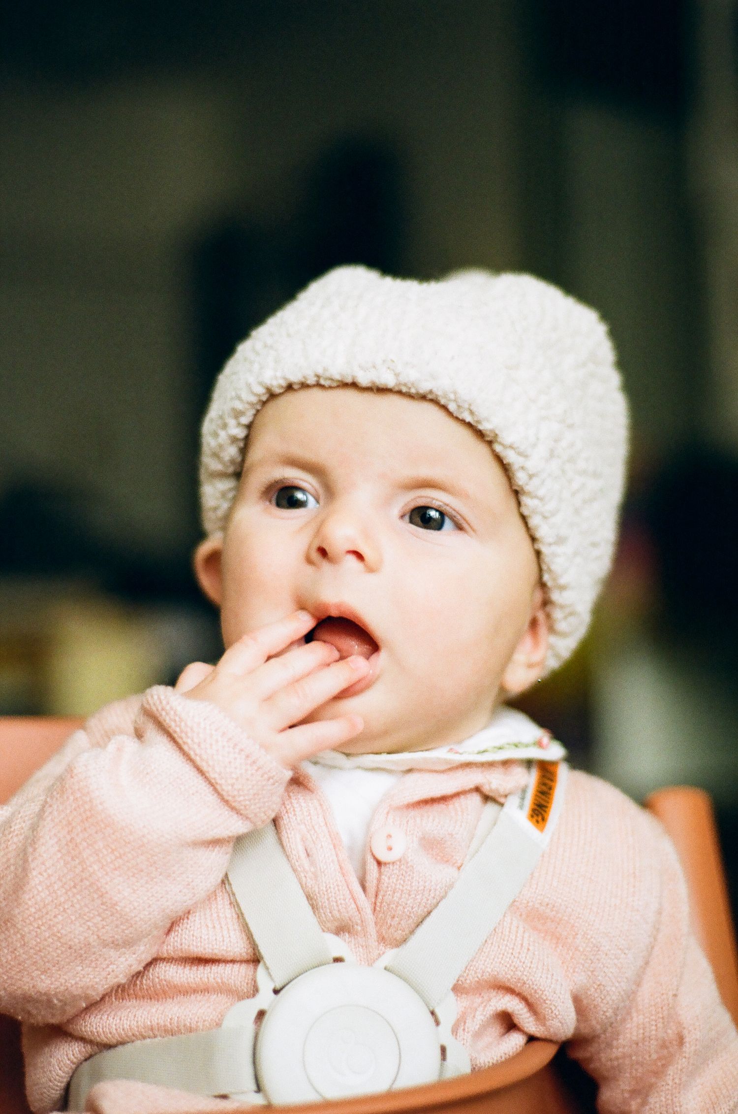 Adorable baby wearing a white fleece hat and pink sweater, hand near mouth.