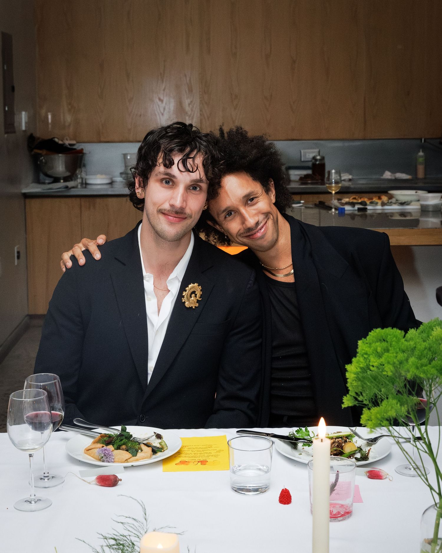 Two men in black blazers smile and pose together at an elegant candlelit dinner table.