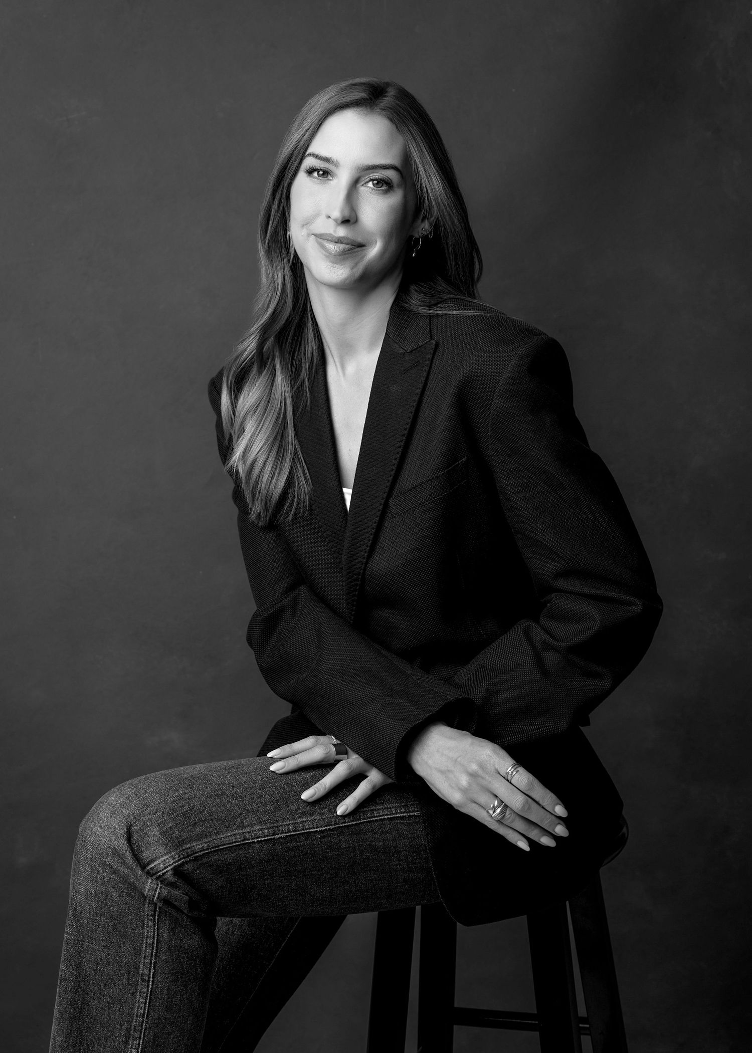 Black and white portrait of woman in blazer and jeans seated on stool.