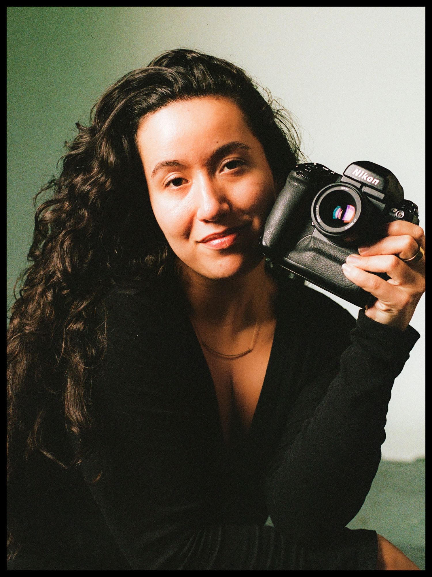 Young woman with curly hair smiles while holding a Nikon camera in a studio.