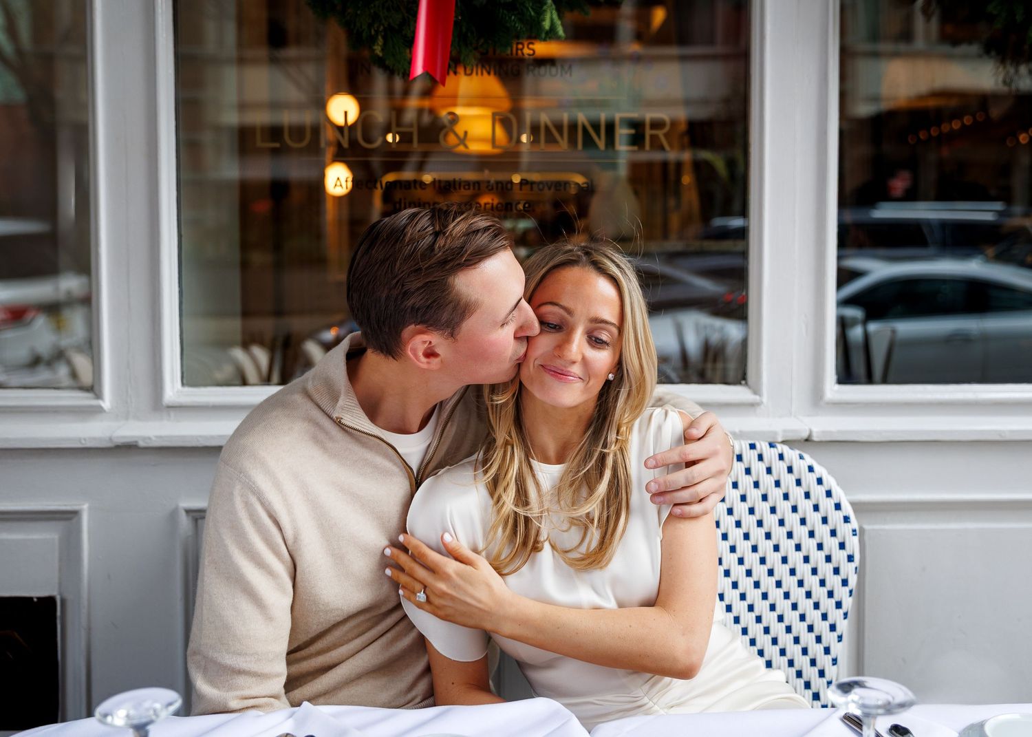 Couple at outdoor cafe table, man kissing woman on cheek as she holds a drink.