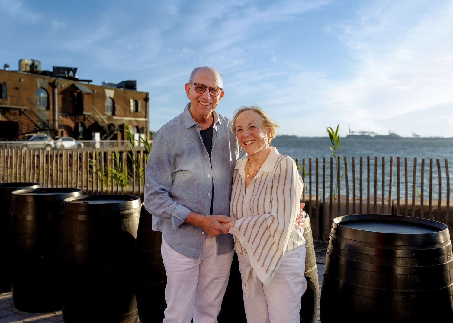 Smiling older couple poses outdoors on a sunny terrace with ocean views behind them.