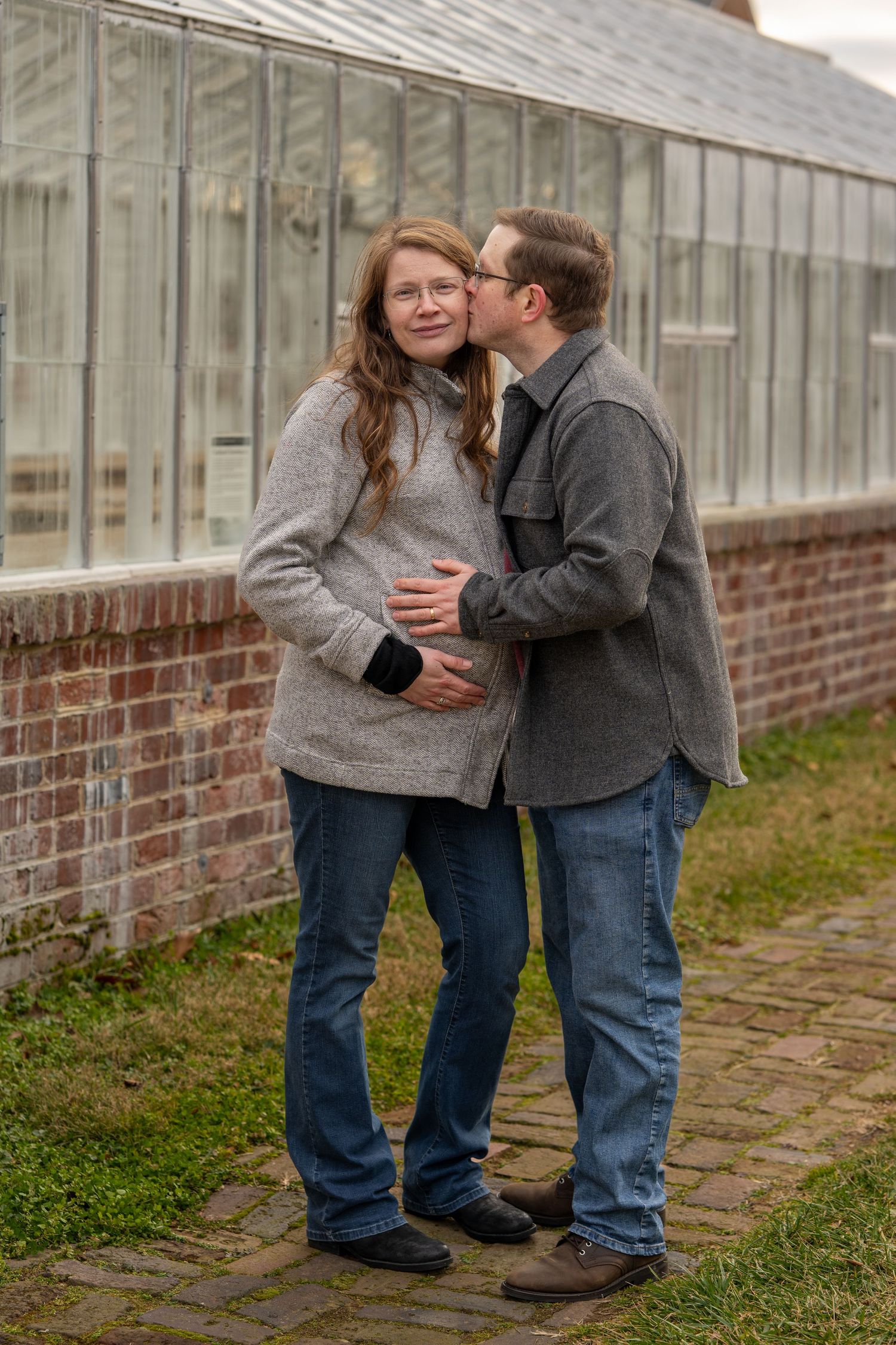 Sweet Maternity photo Chatham Fredericksburg Virginia  in front of green house both have hands on belly and Dad is kissing Mom on cheek.