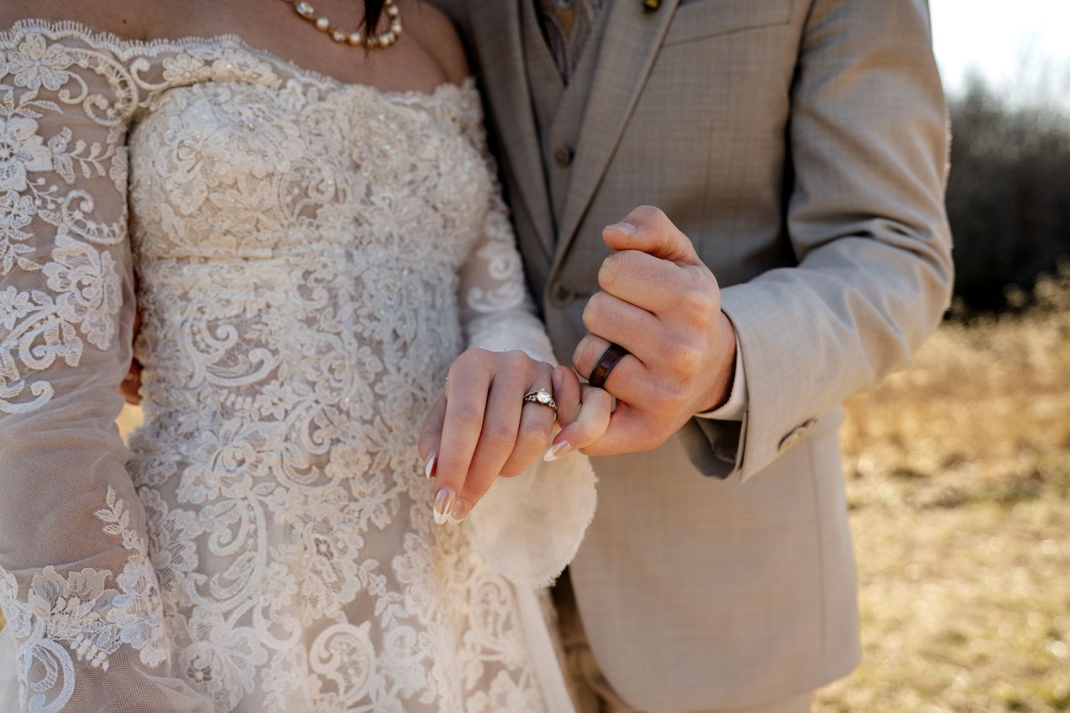 Close-up of a bride and groom linking pinky fingers showing their wedding rings, bride in a lace off-the-shoulder gown groom in a light suit