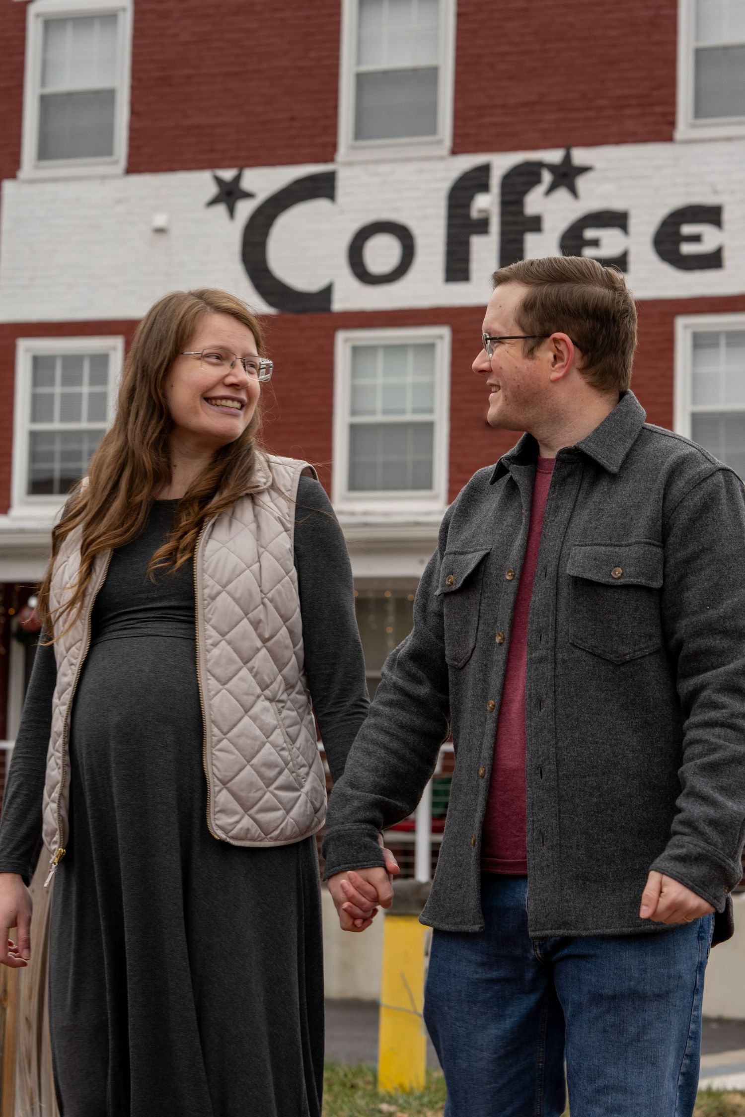 Maternity photo mom and dad holding hands walking in front of brick building with the word Coffee on it. Fredericksburg, Virginia