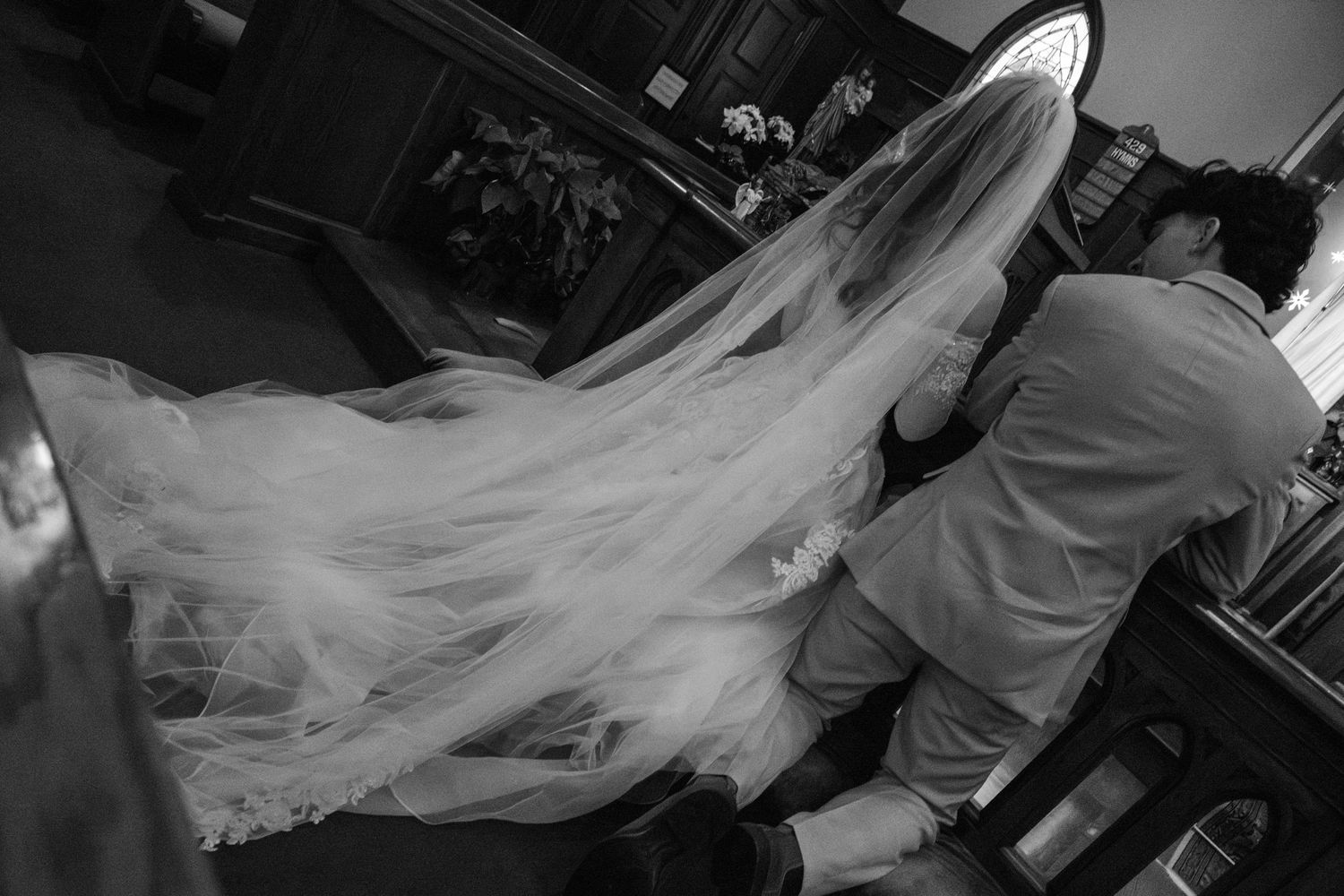 Ethereal black and white of bride and groom kneeling at altar rail praying. Veil spread out behind bride.