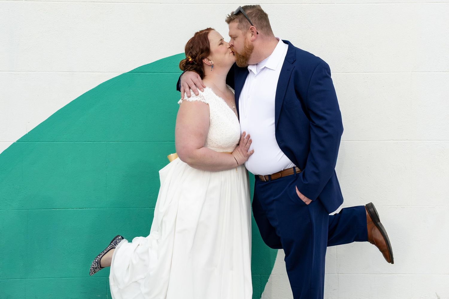 Bride and Groom kissing feet kicked up in front of a white wall with teal pop art painted on it. Richmond, Virginia