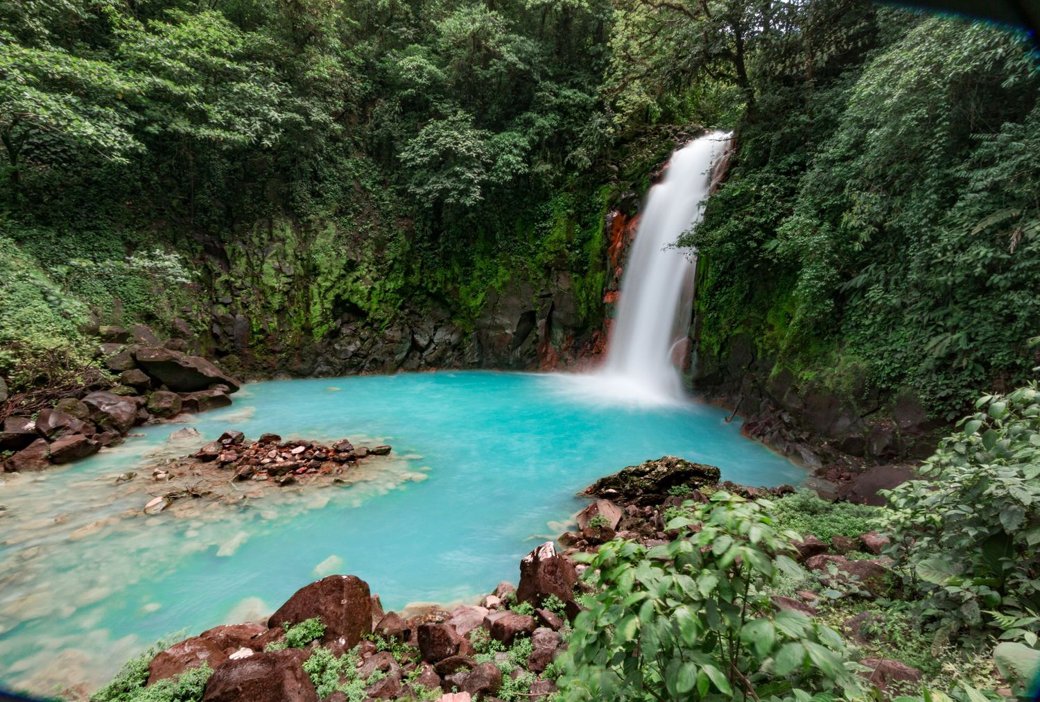 Turquoise blue waterfall cascades through lush green tropical forest landscape.