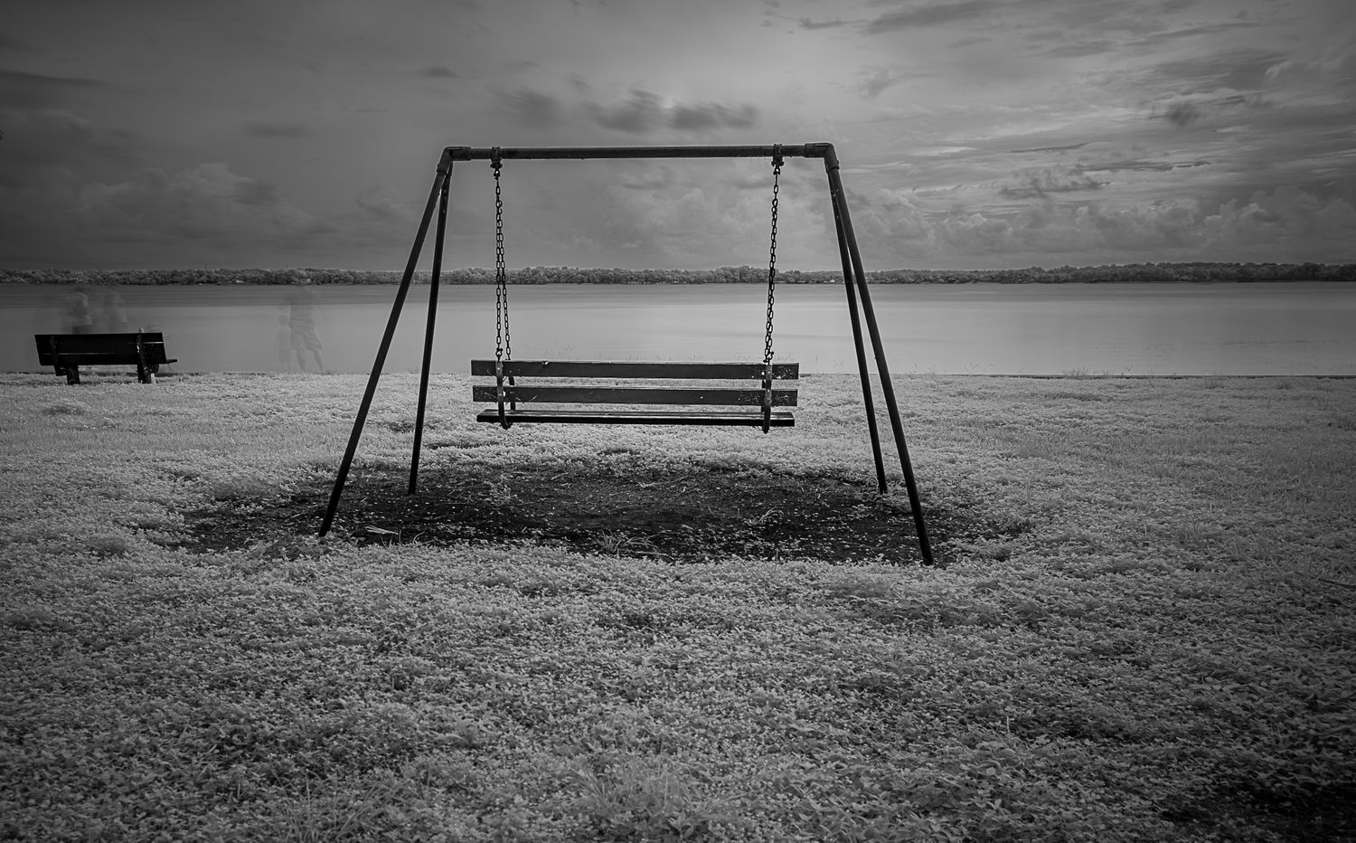 Black and white photo of an empty swing set on a frosty field near a calm lake under an overcast sky.