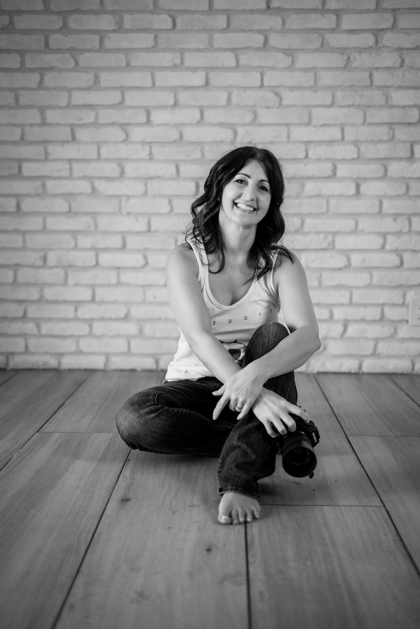 Black and white photo of a smiling woman sitting cross-legged on a wooden floor holding a camera against a brick wall.