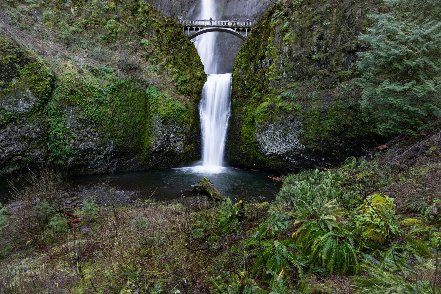 Mossy green waterfall cascading over rocks beneath an arched bridge in a lush forest setting.
