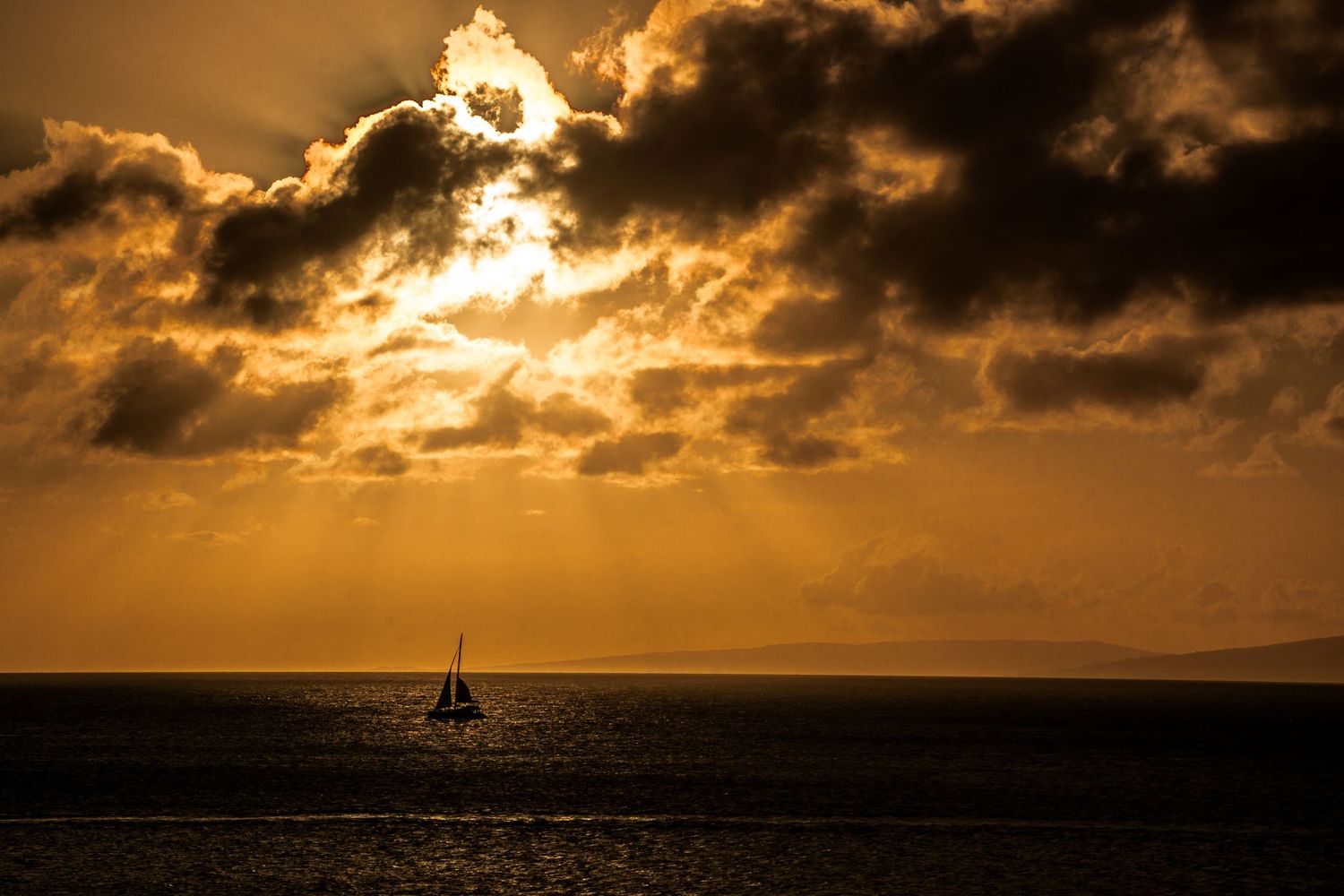 Golden sunset silhouettes sailboat on horizon with dramatic cloudy sky.