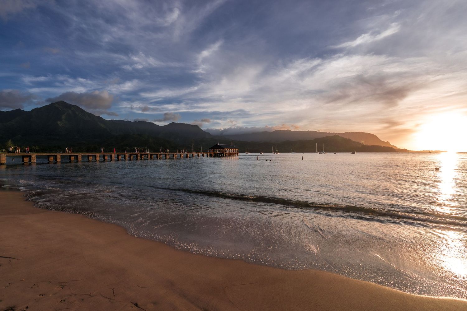 Panoramic beach landscape with mountains and pier silhouetted against a colorful evening sky.
