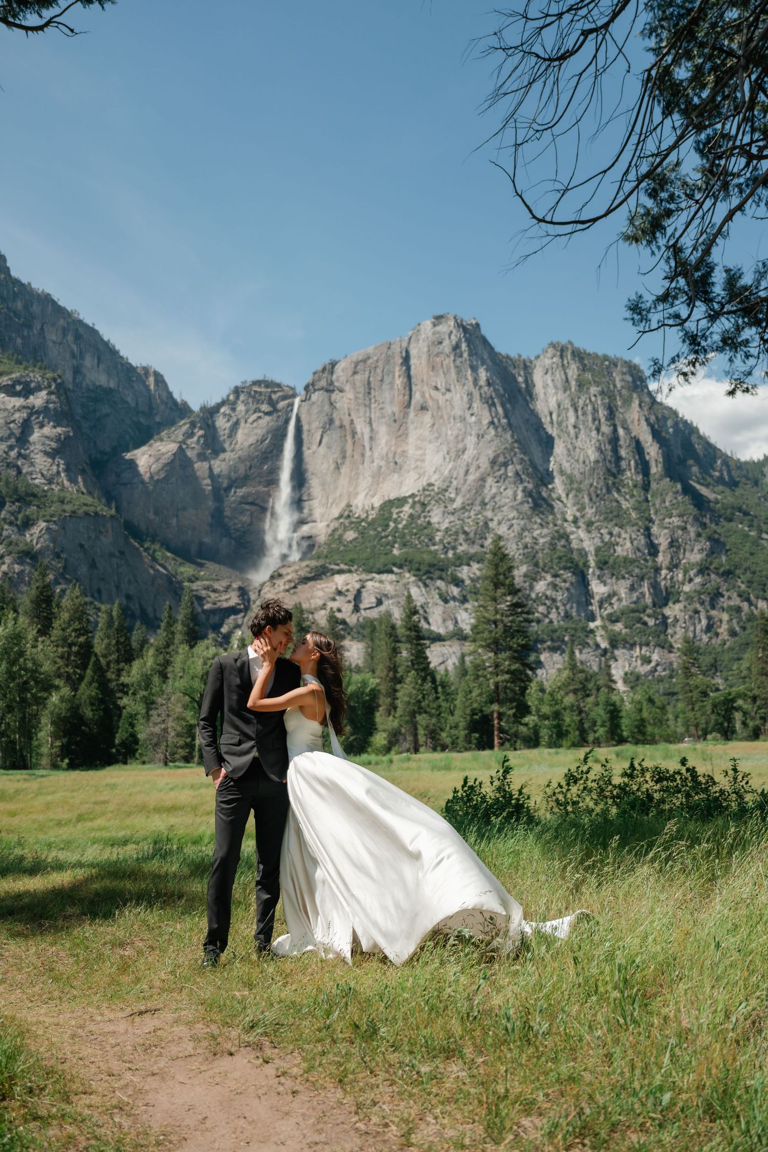 Bride and groom kissing in Yosemite Valley meadow with Yosemite Falls and granite cliffs backdrop.
