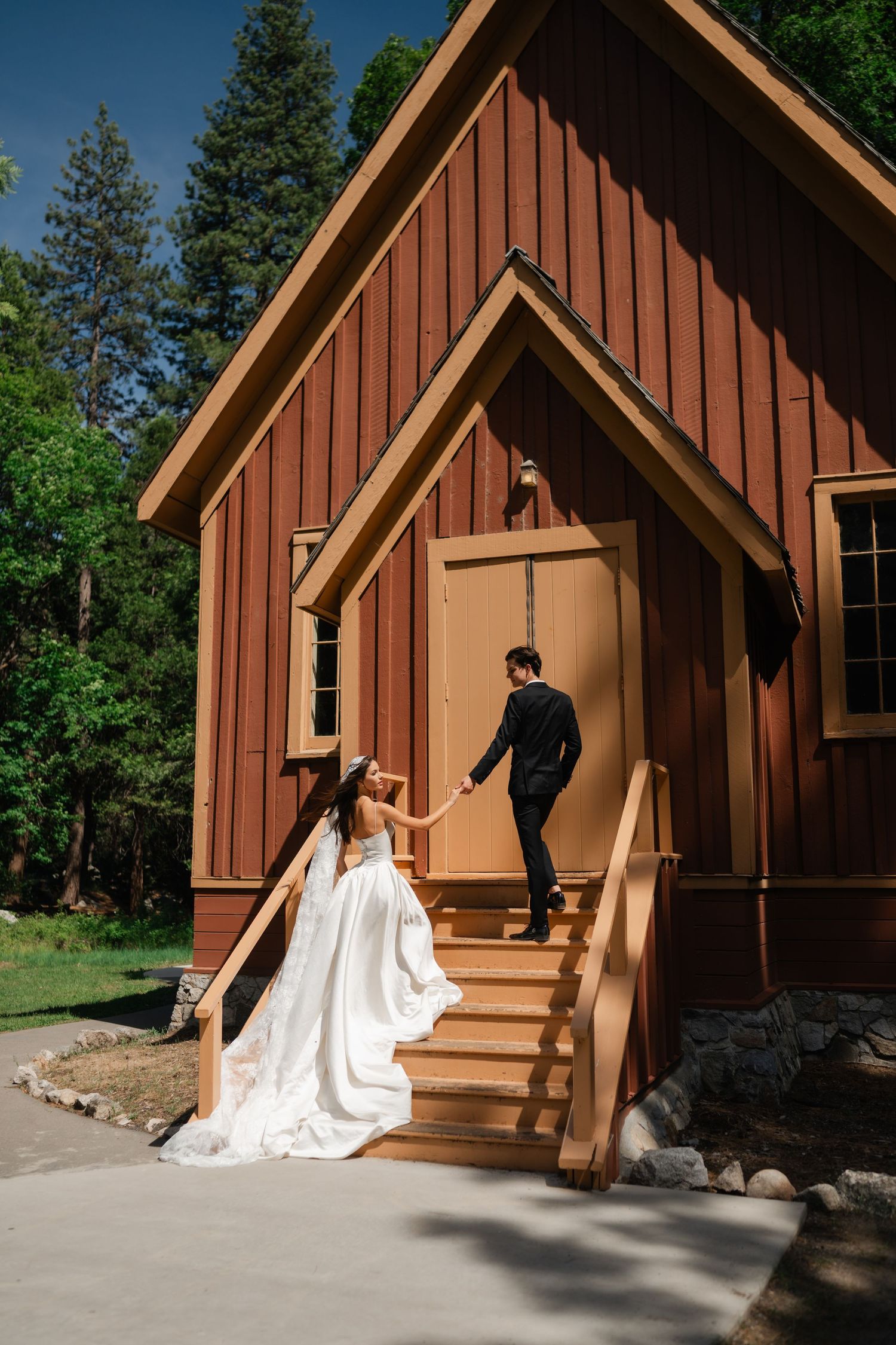 Bride in white gown and groom in black suit hold hands on steps of rustic red chapel surrounded by tall trees.