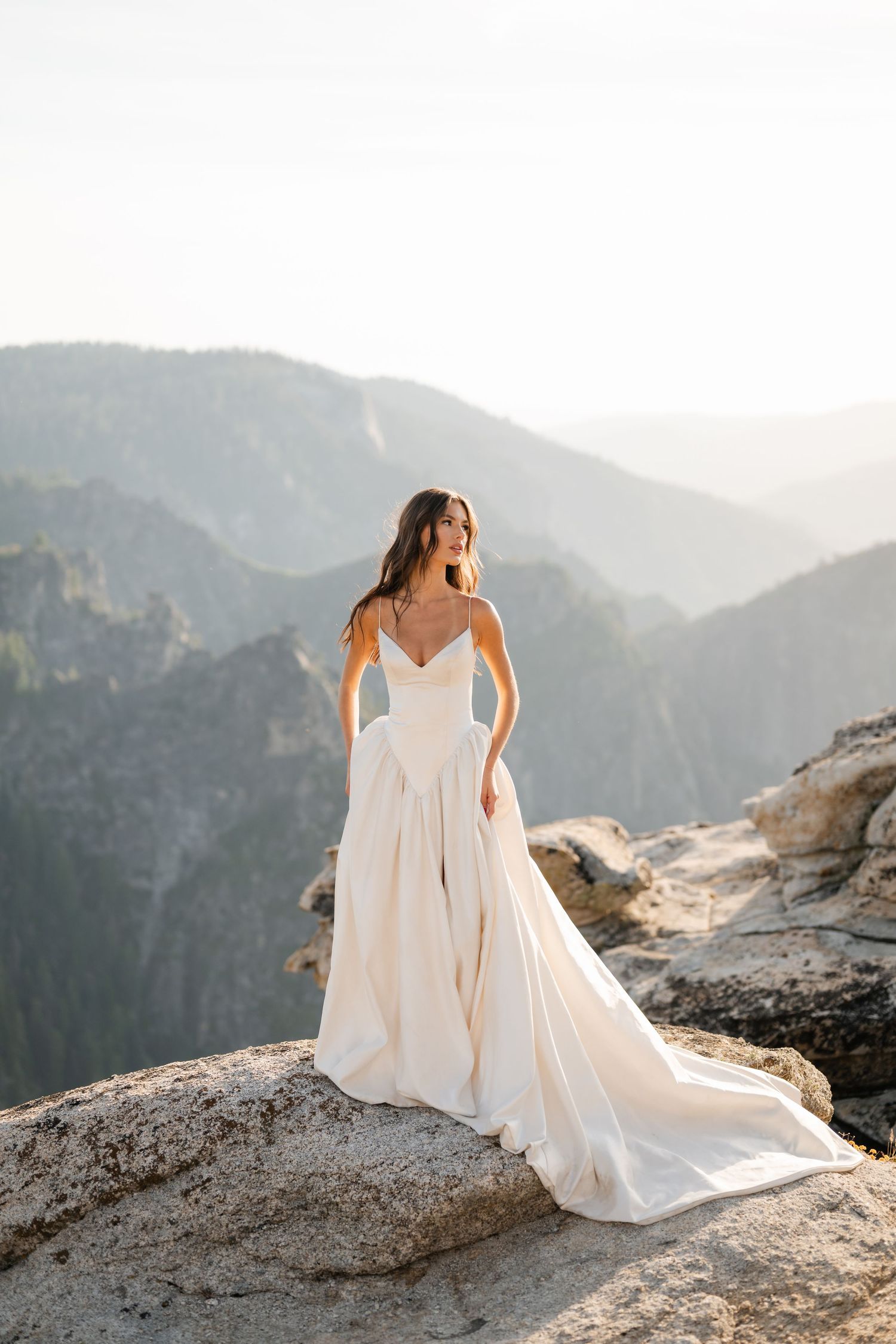 Bride in flowing ivory spaghetti-strap wedding gown poses on rocky mountain overlook at golden hour.