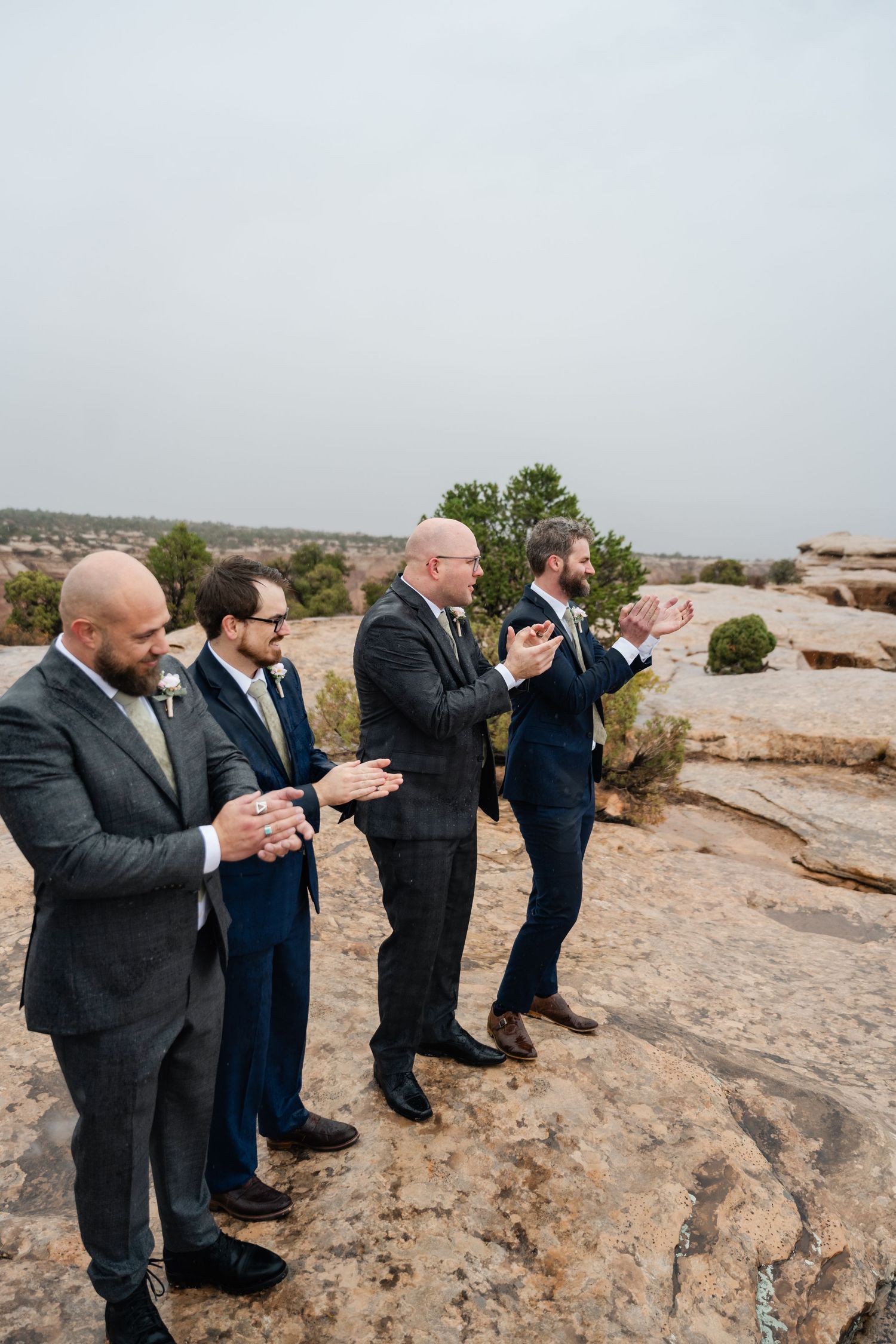 People in dark suits stand in a line on rocky terrain, clapping and celebrating.