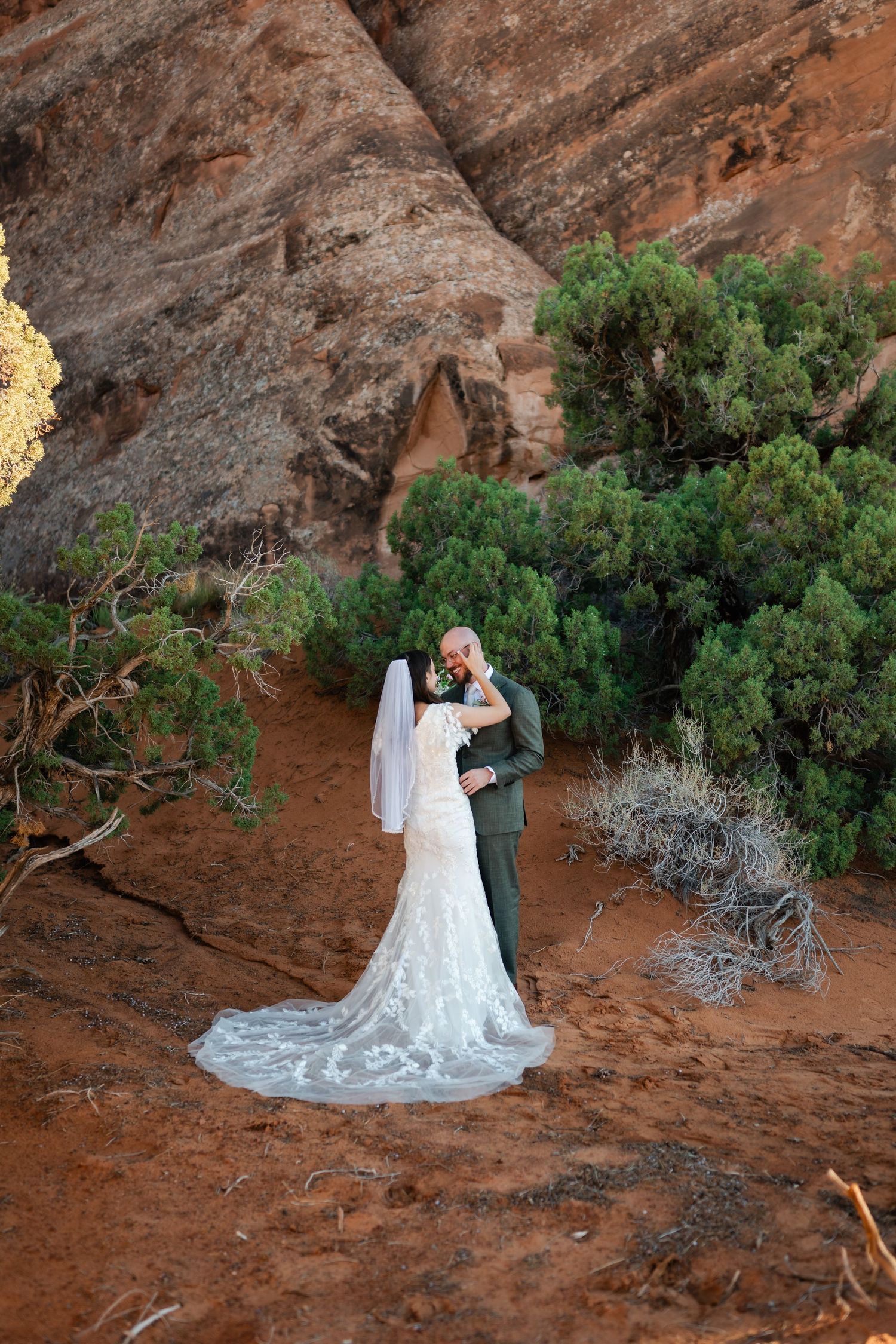 A wedding couple poses against red rock formations and desert brush in a scenic outdoor setting.