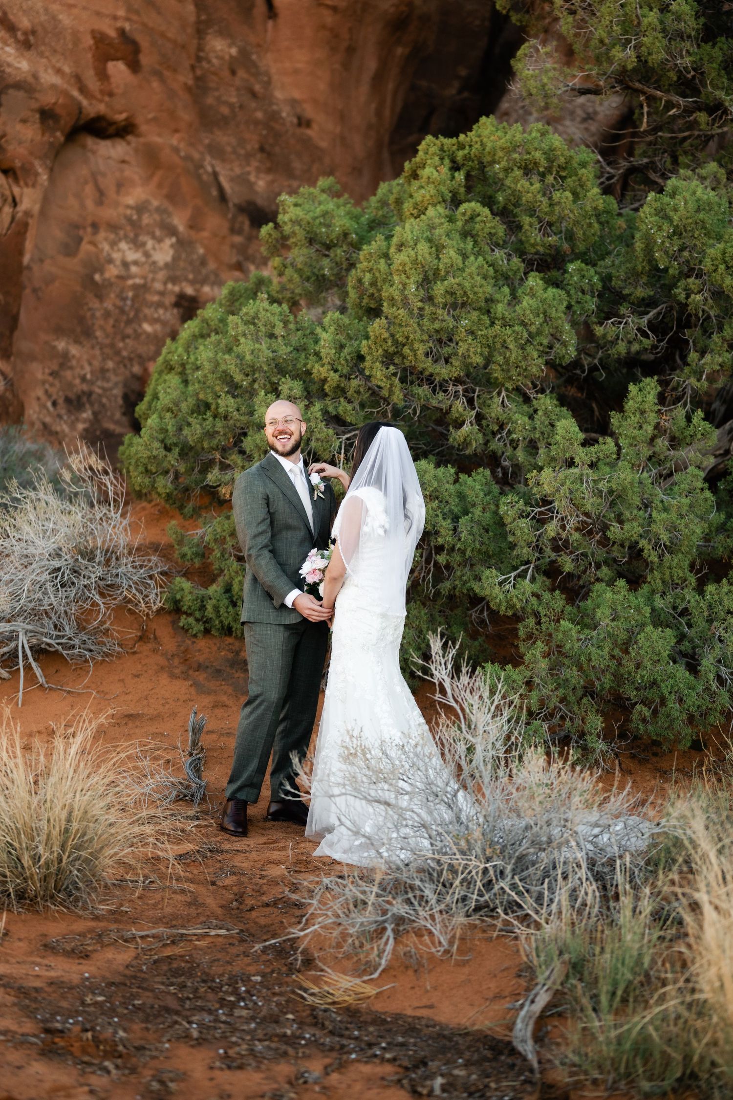 A couple poses for wedding photos against rustic red rocks and desert vegetation.