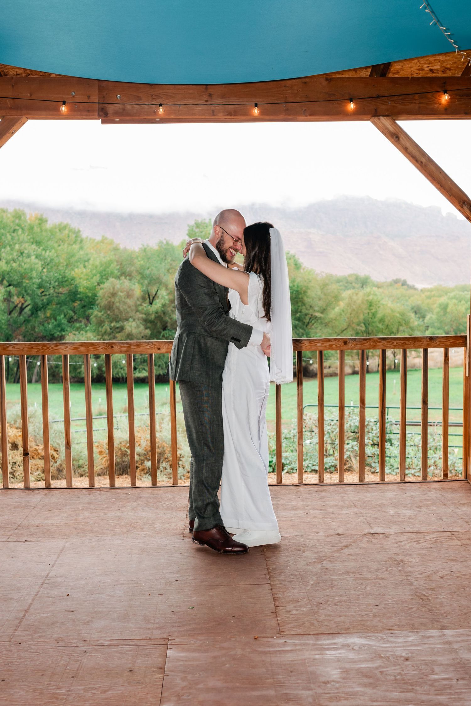 A couple embracing on a covered deck with mountain views and string lights overhead.
