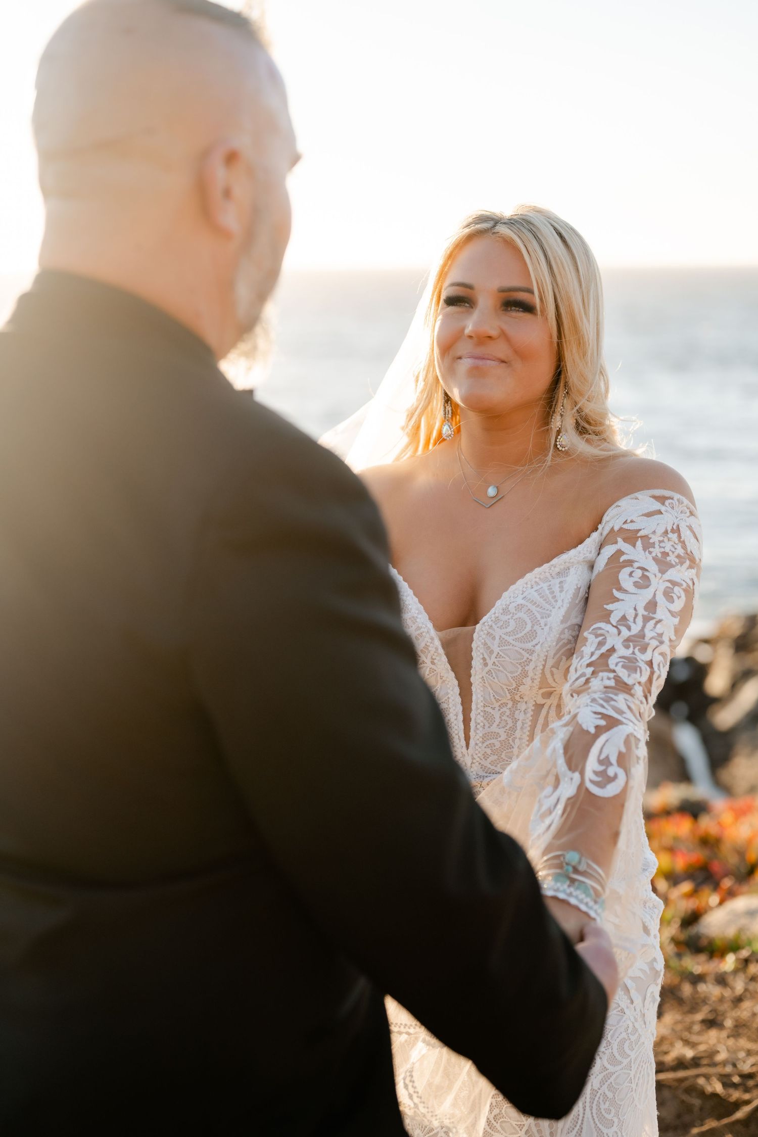 A romantic wedding ceremony takes place on a rocky coastal beach during golden hour sunset.