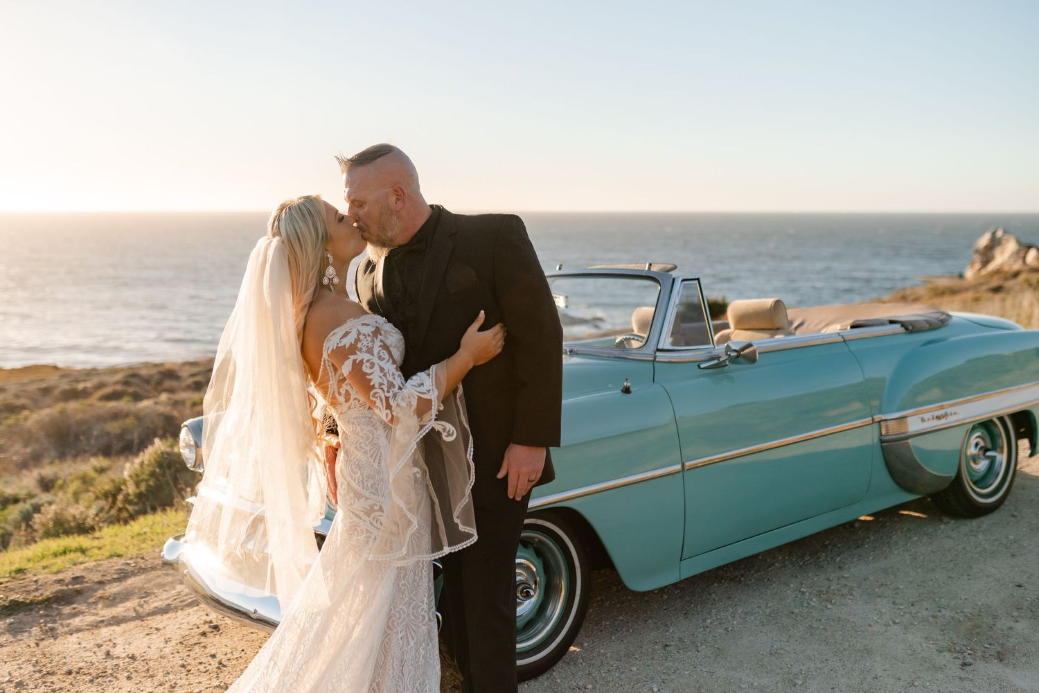 Newlyweds pose by vintage turquoise car against coastal backdrop at sunset.