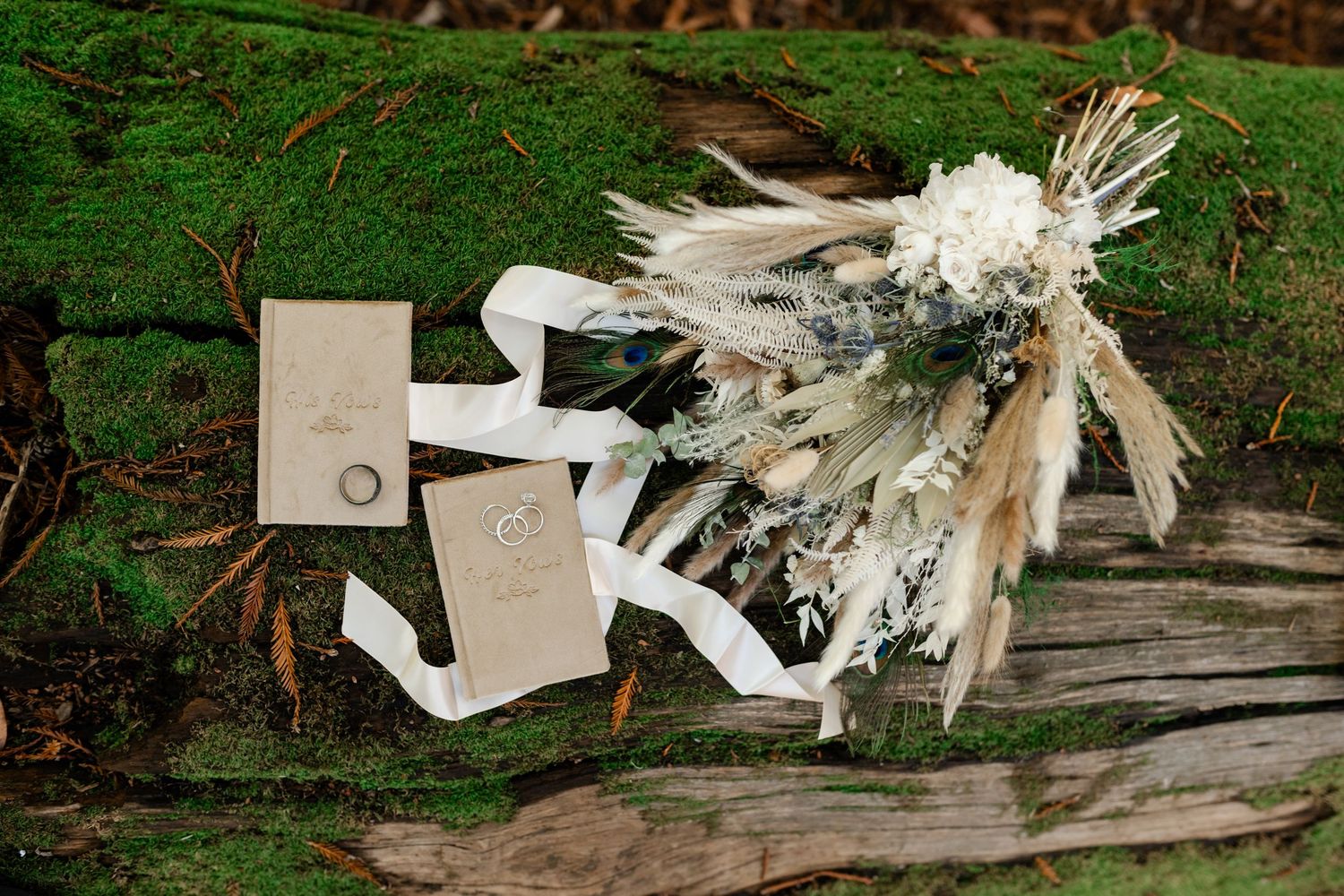 White wedding shoes and a dried floral bouquet with pampas grass rest on a mossy log in an outdoor setting.