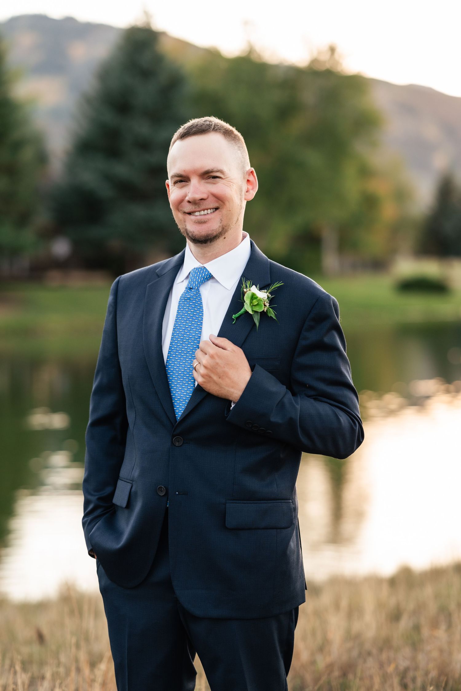Groom in navy suit and blue tie poses for formal portraits near a lake at sunset.