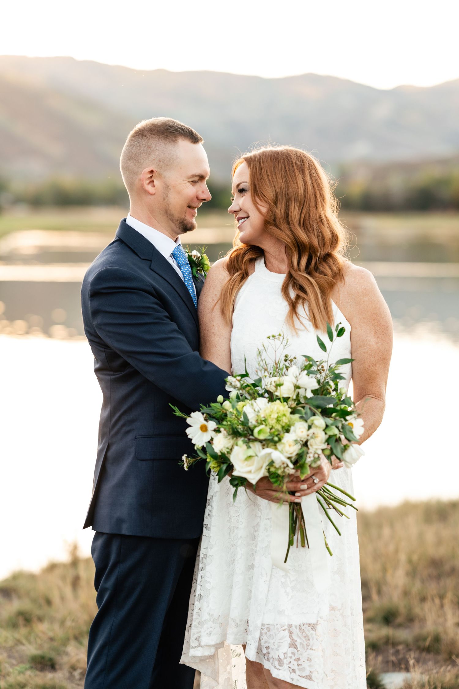 A couple shares a romantic moment outdoors with a beautiful white floral bouquet against a mountain landscape.