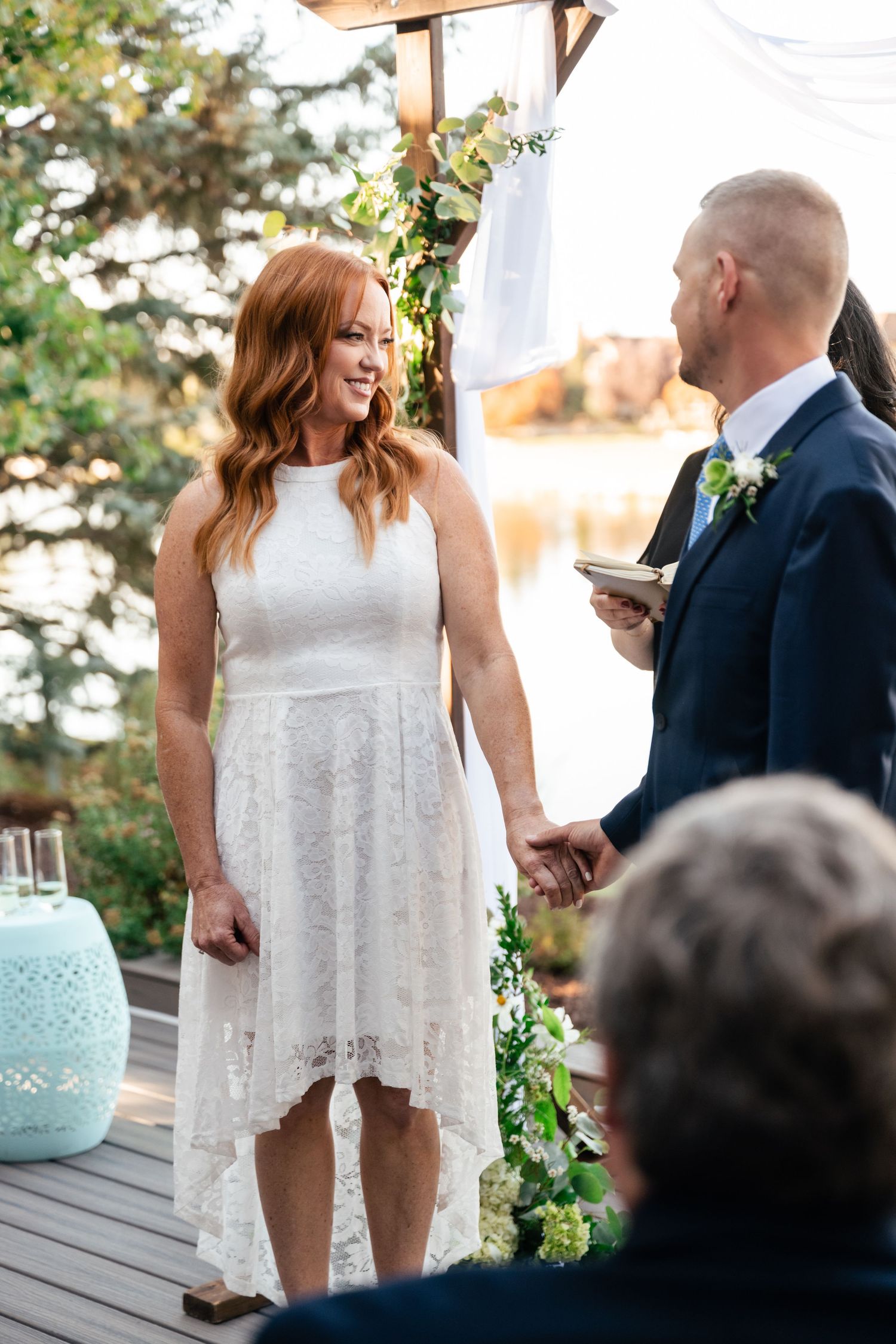 A couple exchanges vows under a wooden arch decorated with white fabric and greenery.