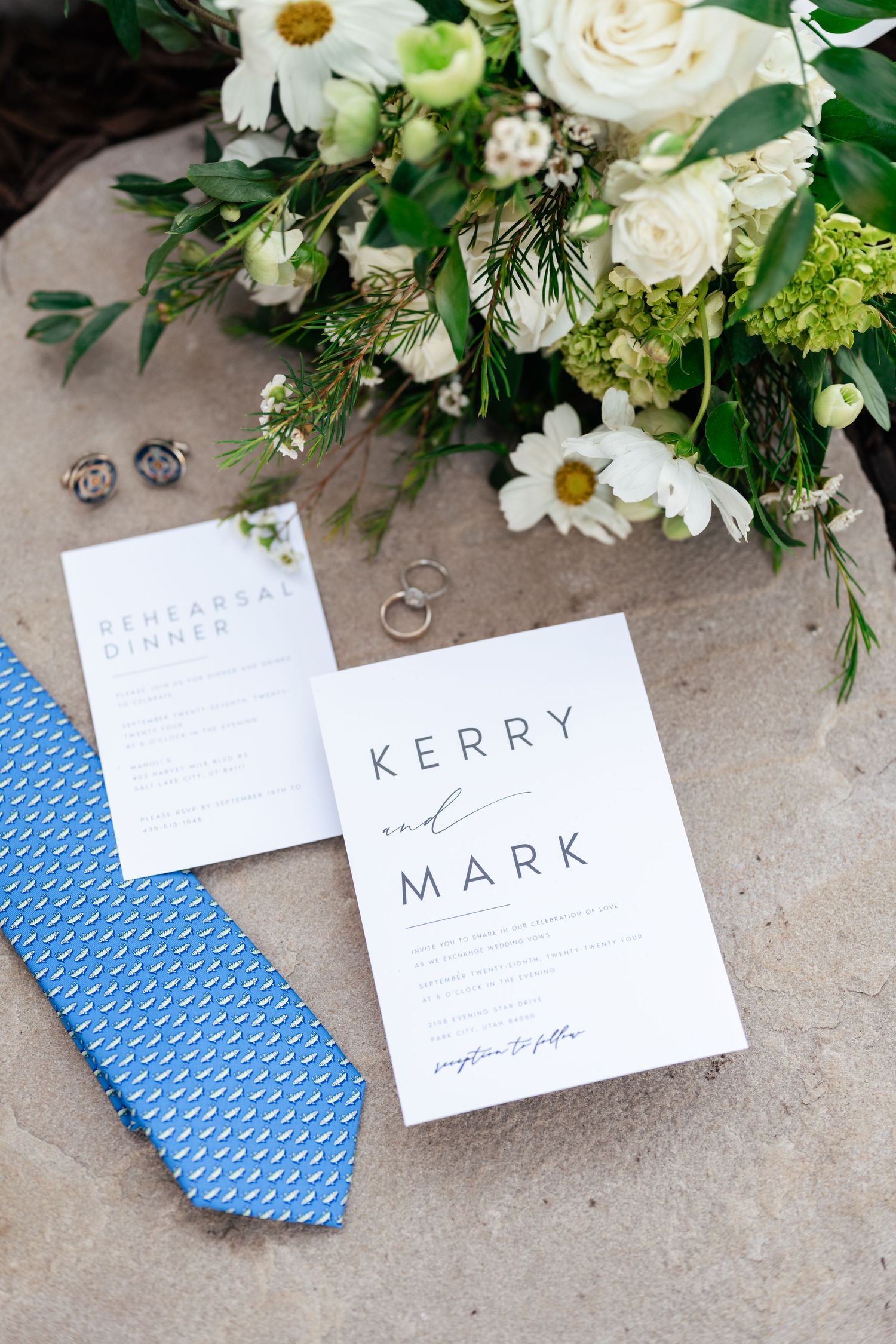 Wedding invitation suite with white flowers and blue tie displayed on grey stone surface.